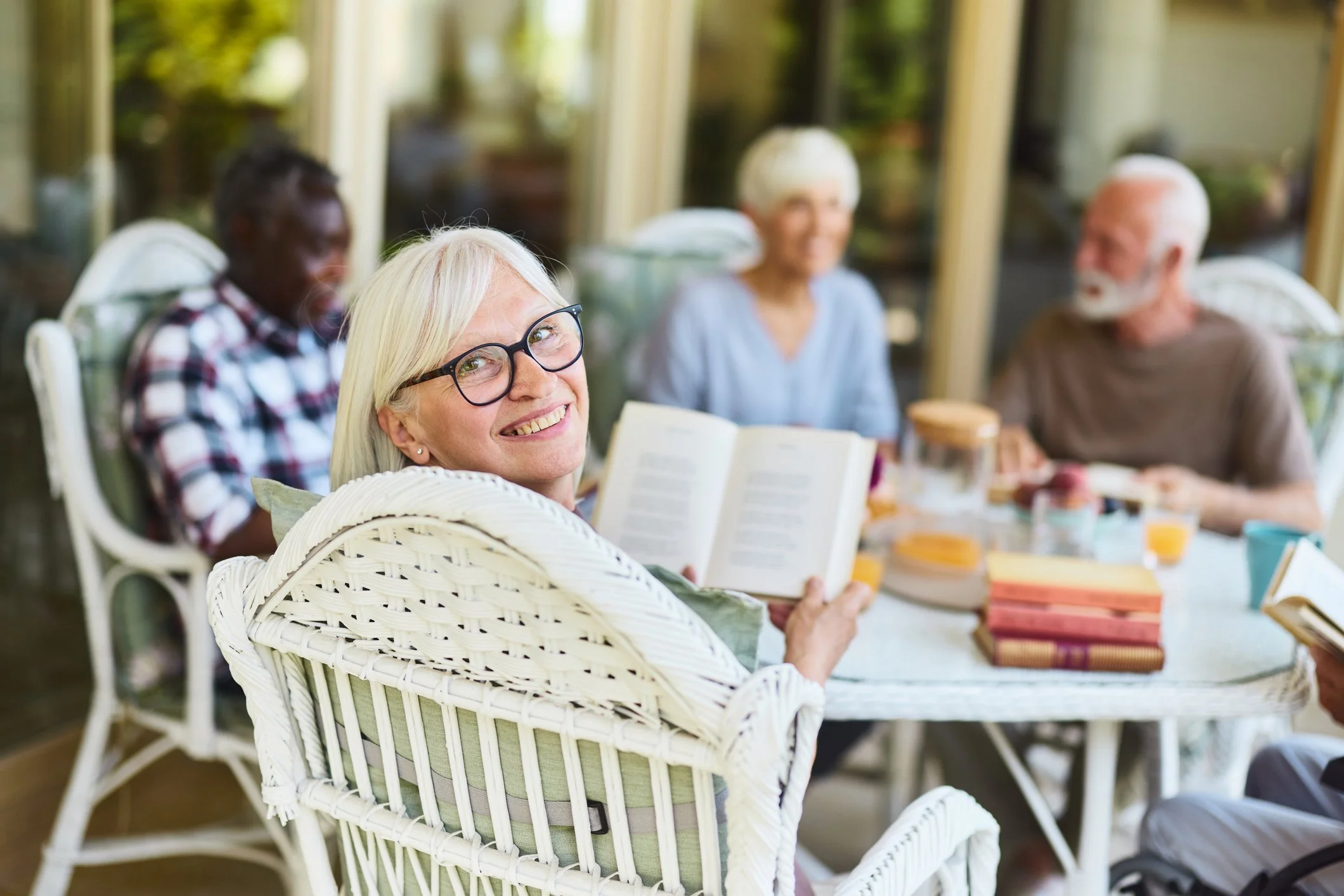 A group of five older adults in a cozy outdoor setting, sitting around a table with books, beverages, and snacks, enjoying a pleasant conversation.