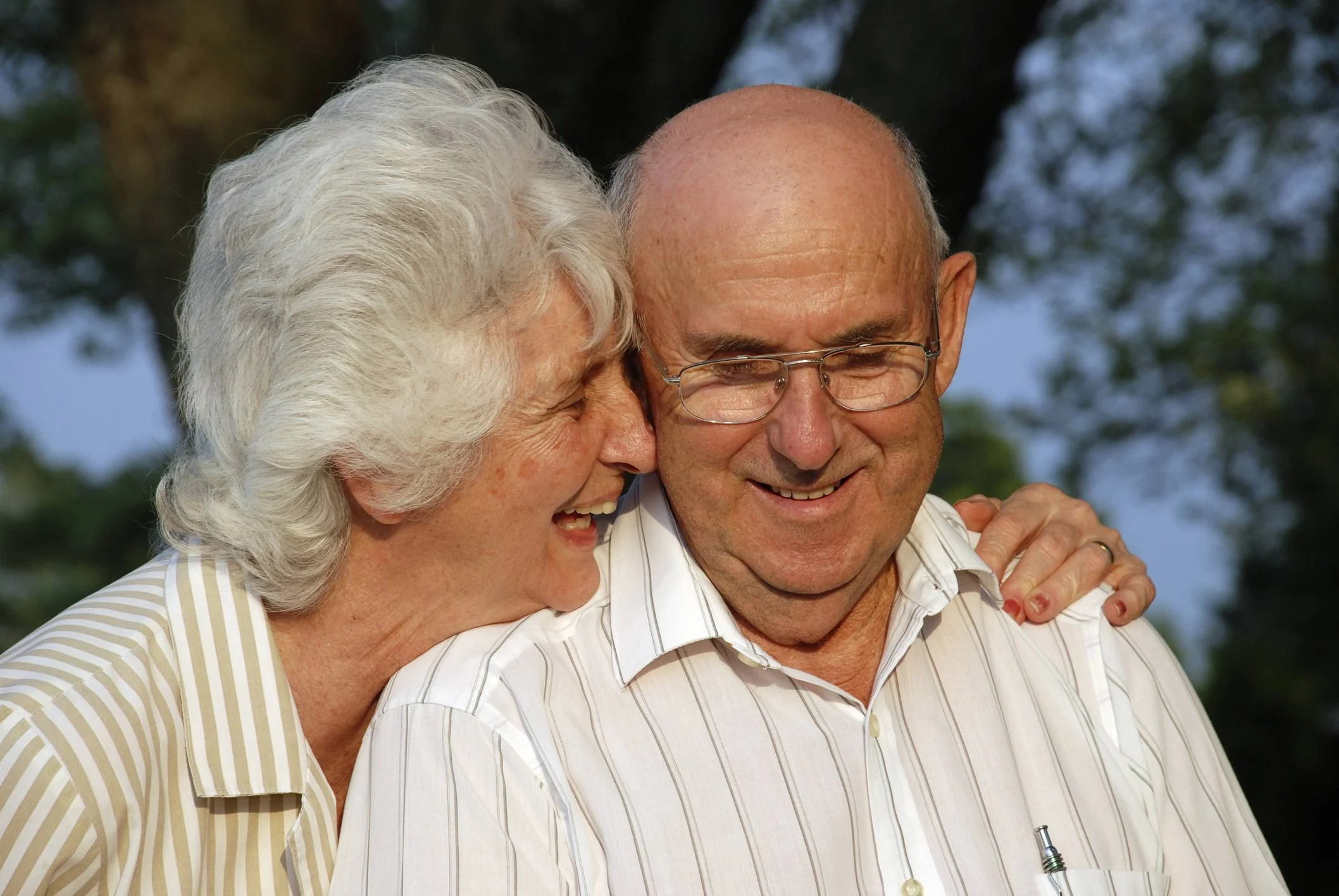 An elderly woman is smiling and whispering into the ear of an elderly man, who is also smiling. They are outdoors, with trees in the background, and the woman has her hand on the man's shoulder.