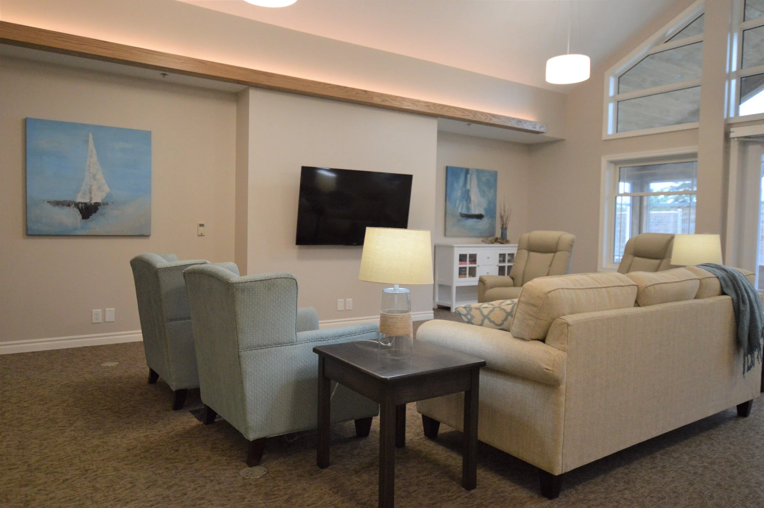 Living room with beige sofa and armchairs, wooden coffee table with a lamp, wall-mounted TV, large windows, nautical-themed artwork, white cabinet, and a ceiling light.