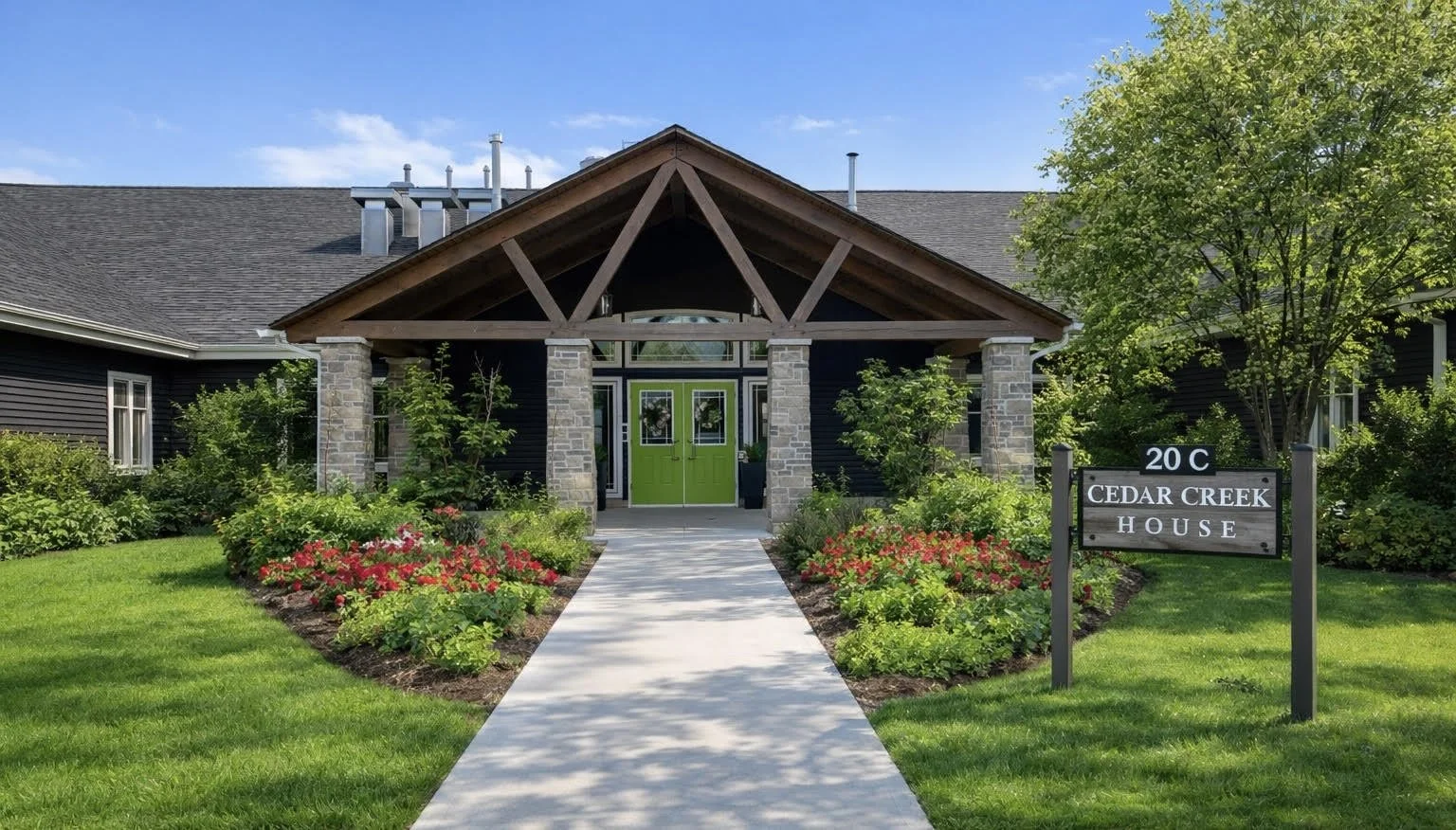 Front view of a house named Cedar Creek House, with a green front door, stone columns, and a gabled roof, surrounded by a landscaped garden with flowers and a lawn.