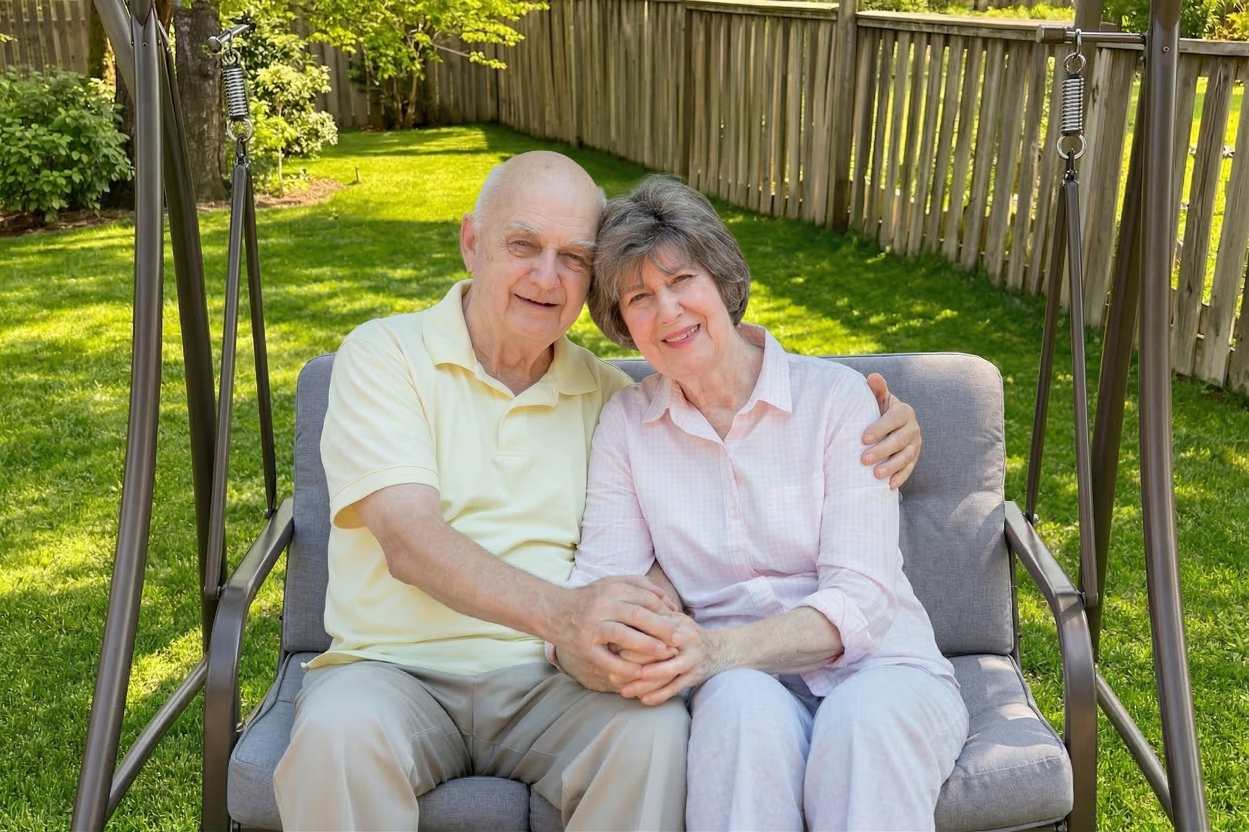 An elderly couple sitting on a gray outdoor swing in their backyard, smiling and holding hands, with a green lawn and wooden fence in the background.