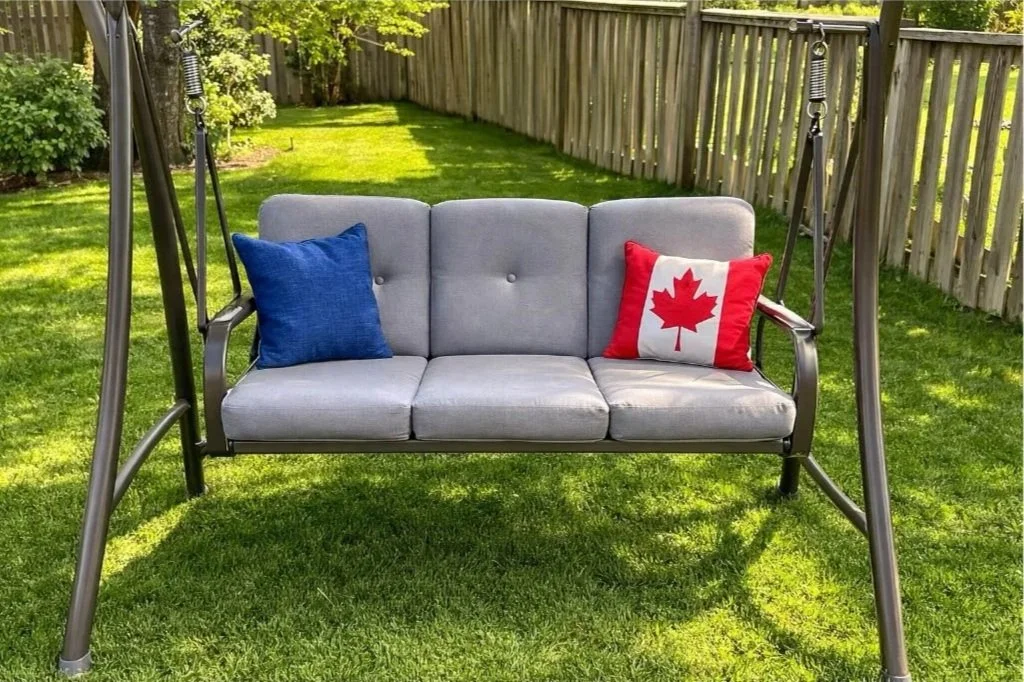 A gray outdoor bench swing with two decorative pillows, one blue and one with a Canadian flag design, in a fenced backyard with green grass and trees.