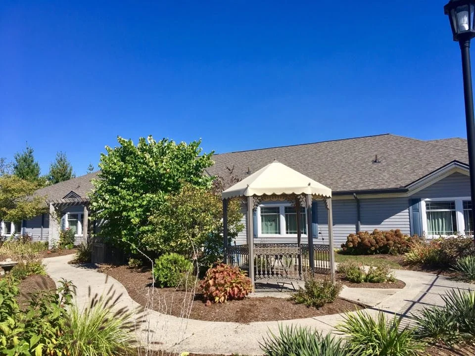 A backyard with a pathway, lush greenery, a gazebo with a white canopy, and a house in the background.
