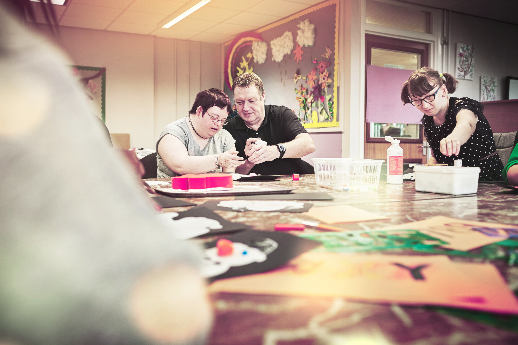 People engaged in a crafting activity at a table in a classroom with colorful decorations on the wall.