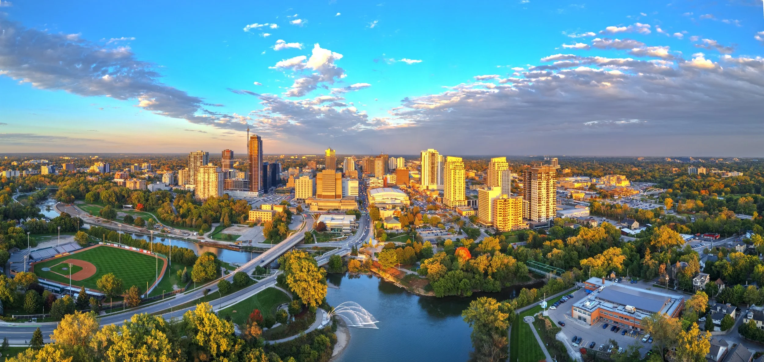 Aerial view of a city skyline at sunset, with tall buildings, a park with a baseball field, a river, and bridges.