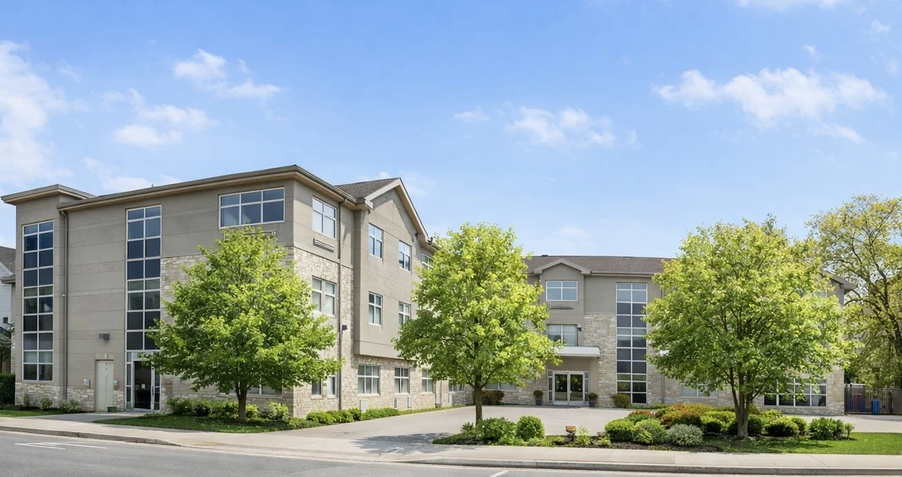 Modern multi-story apartment building with large windows, surrounded by green trees and landscaped bushes, under a blue sky with scattered clouds.
