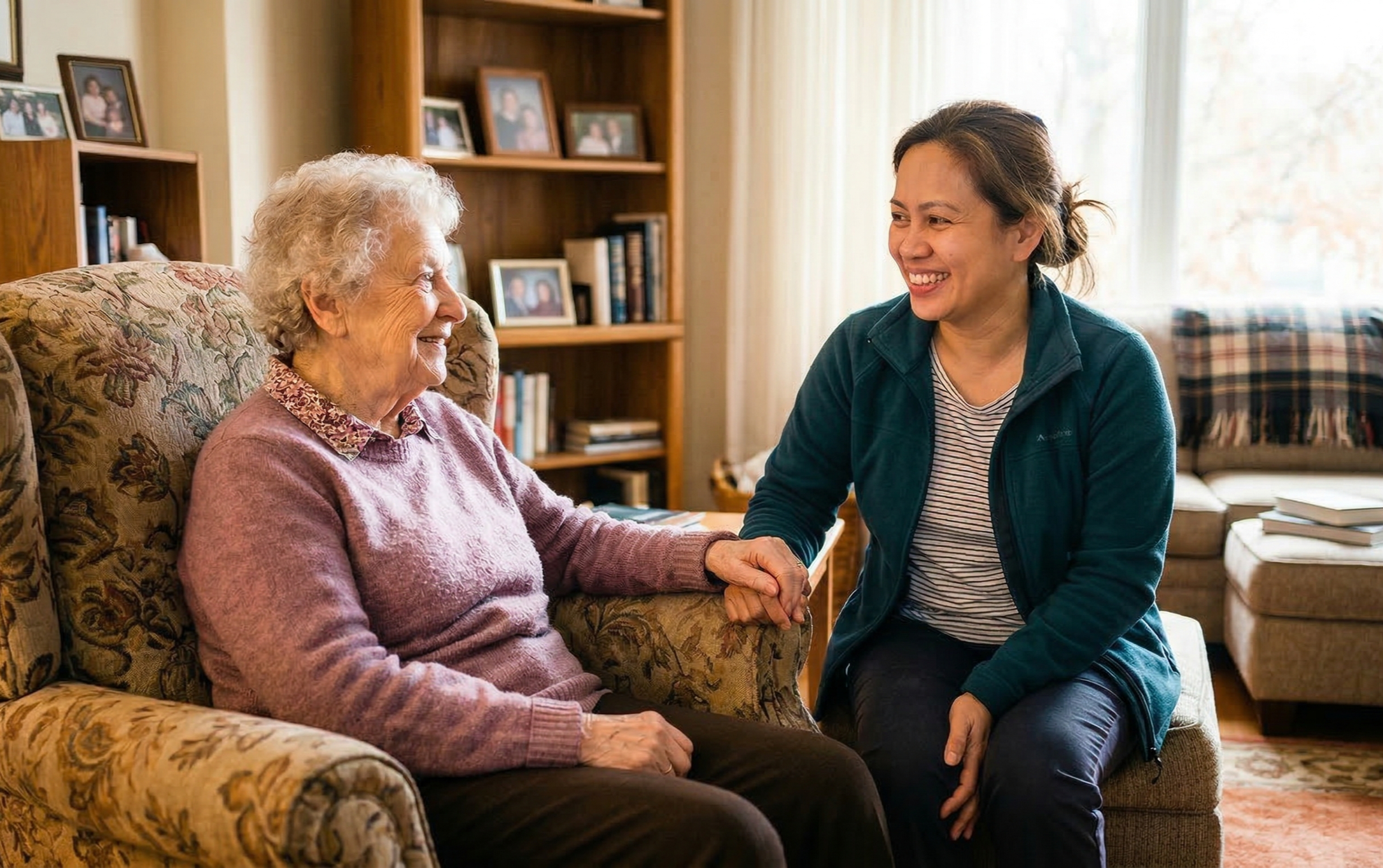 A young woman with brown hair pulled back, wearing a blue jacket and striped shirt, smiles while holding hands with an elderly woman with curly gray hair, dressed in a purple sweater, who is sitting on a floral upholstered armchair in a cozy living room with bookshelves and framed photos in the background.