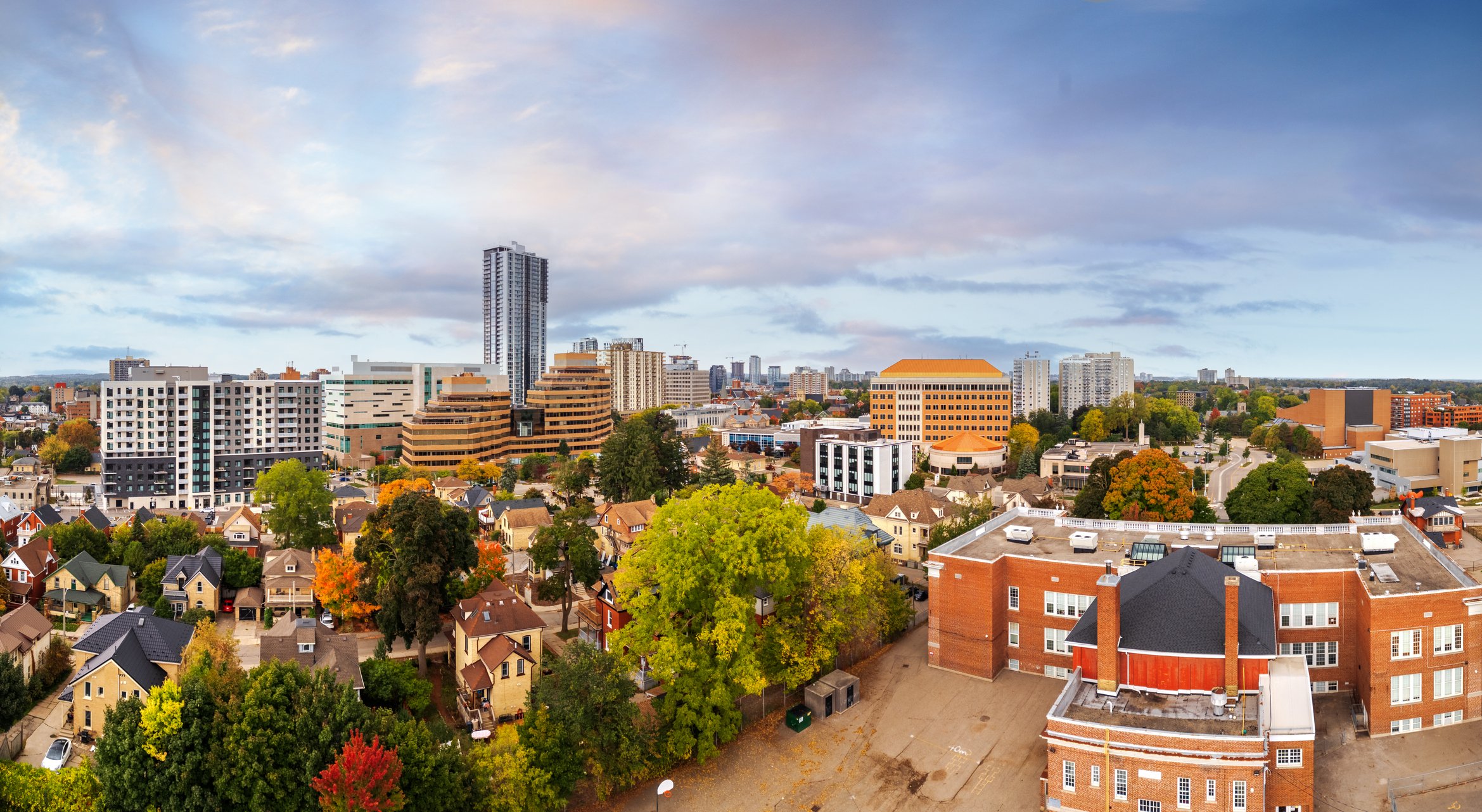 A cityscape with a mix of tall modern buildings and smaller residential houses, with trees showing fall foliage, under a partly cloudy sky.