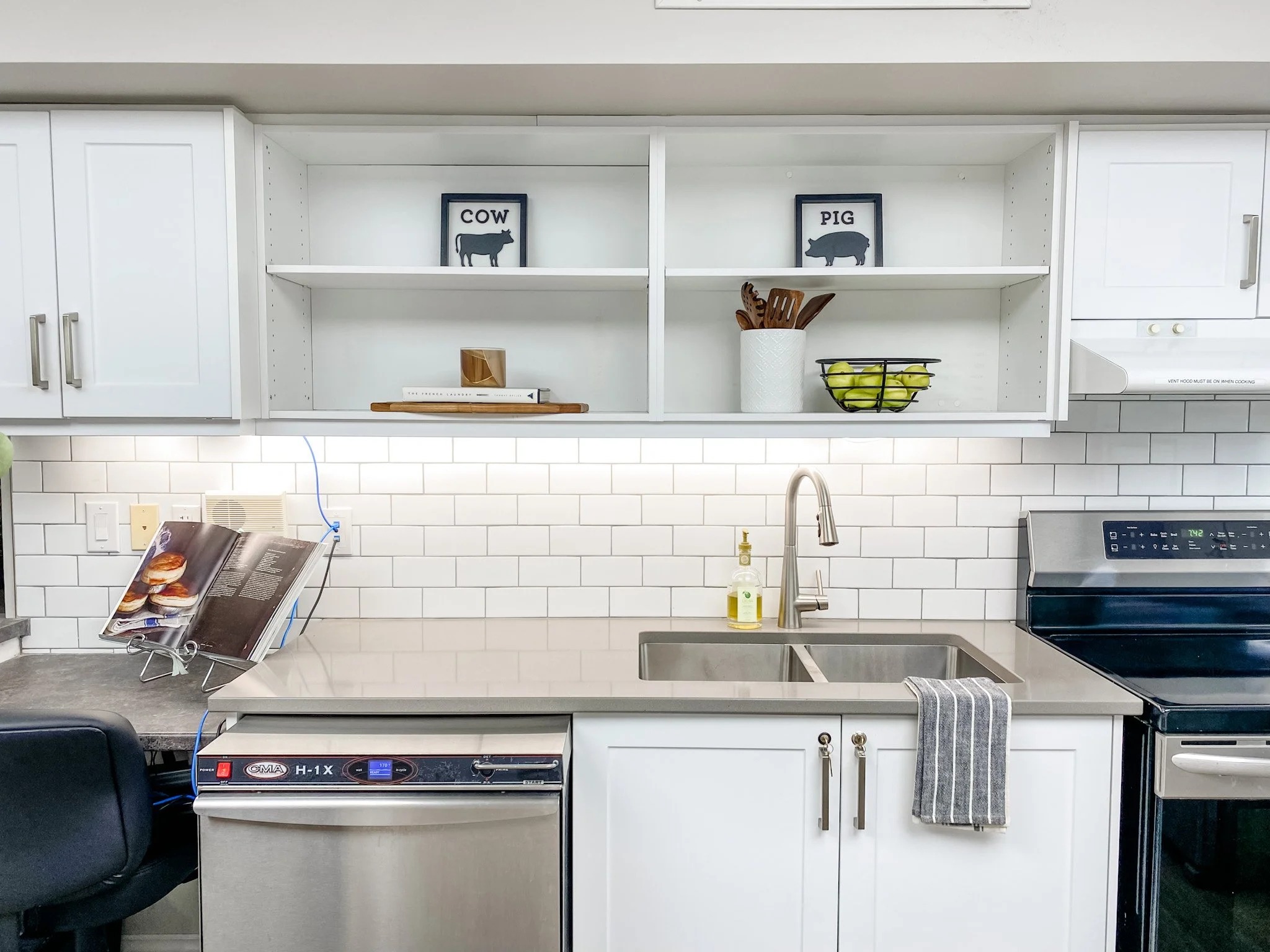 White kitchen with open cabinets, two framed signs labeled 'COW' and 'PIG,' a white backsplash, stainless steel sink, soap dispenser, dish towel, dishwasher, and stove.