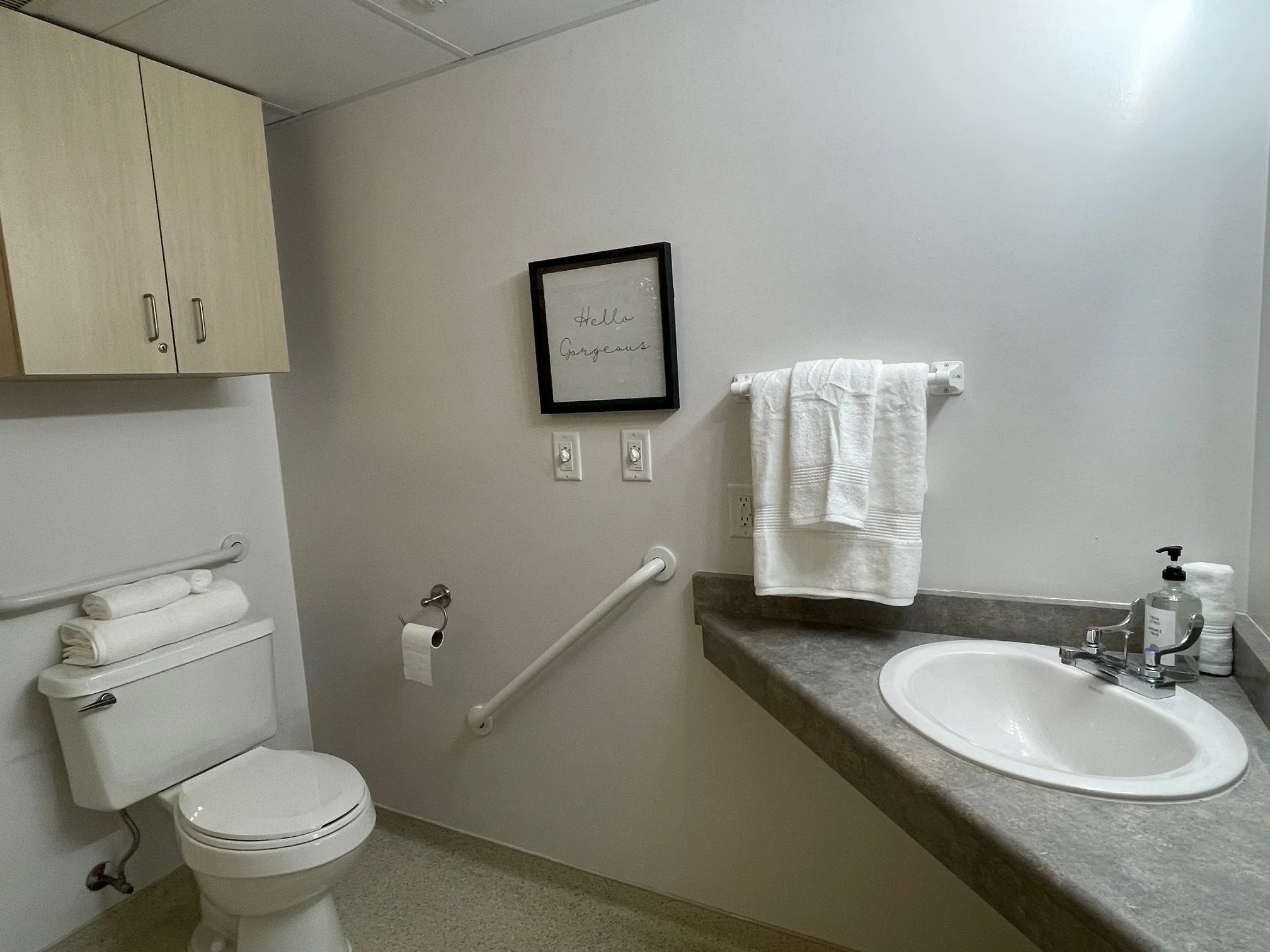 A clean, accessible bathroom with a toilet, a wall-mounted cabinet, a sink with a countertop, a towel rack with a towel, and a black-framed piece of wall art that says 'Hello Gorgeous'.