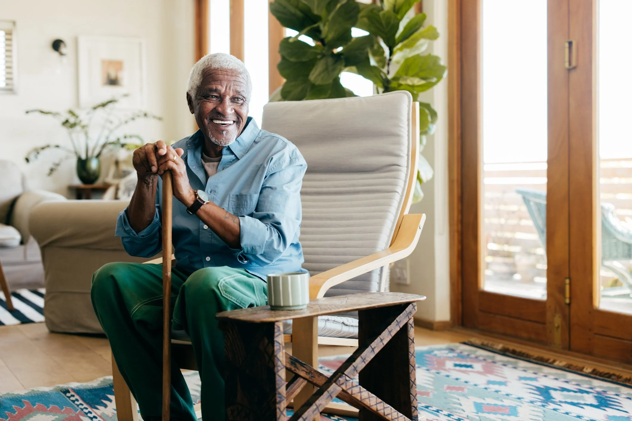 An elderly man with gray hair and a beard smiling while sitting in a modern living room. He is wearing a light blue shirt, green pants, and holding a wooden cane. There is a coffee mug on a small wooden table beside him, and large windows with wooden frames and a sliding door in the background.