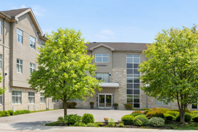 Exterior view of a modern apartment building with large windows, surrounded by green trees and landscaped bushes, on a sunny day.