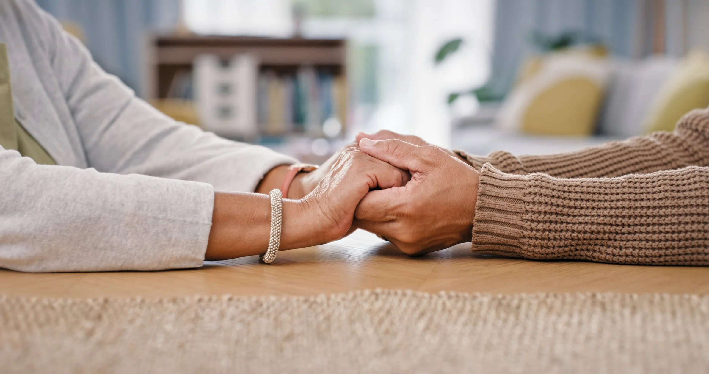 A person gently holding hands with another person across a wooden table, with a cozy, well-lit room in the background.