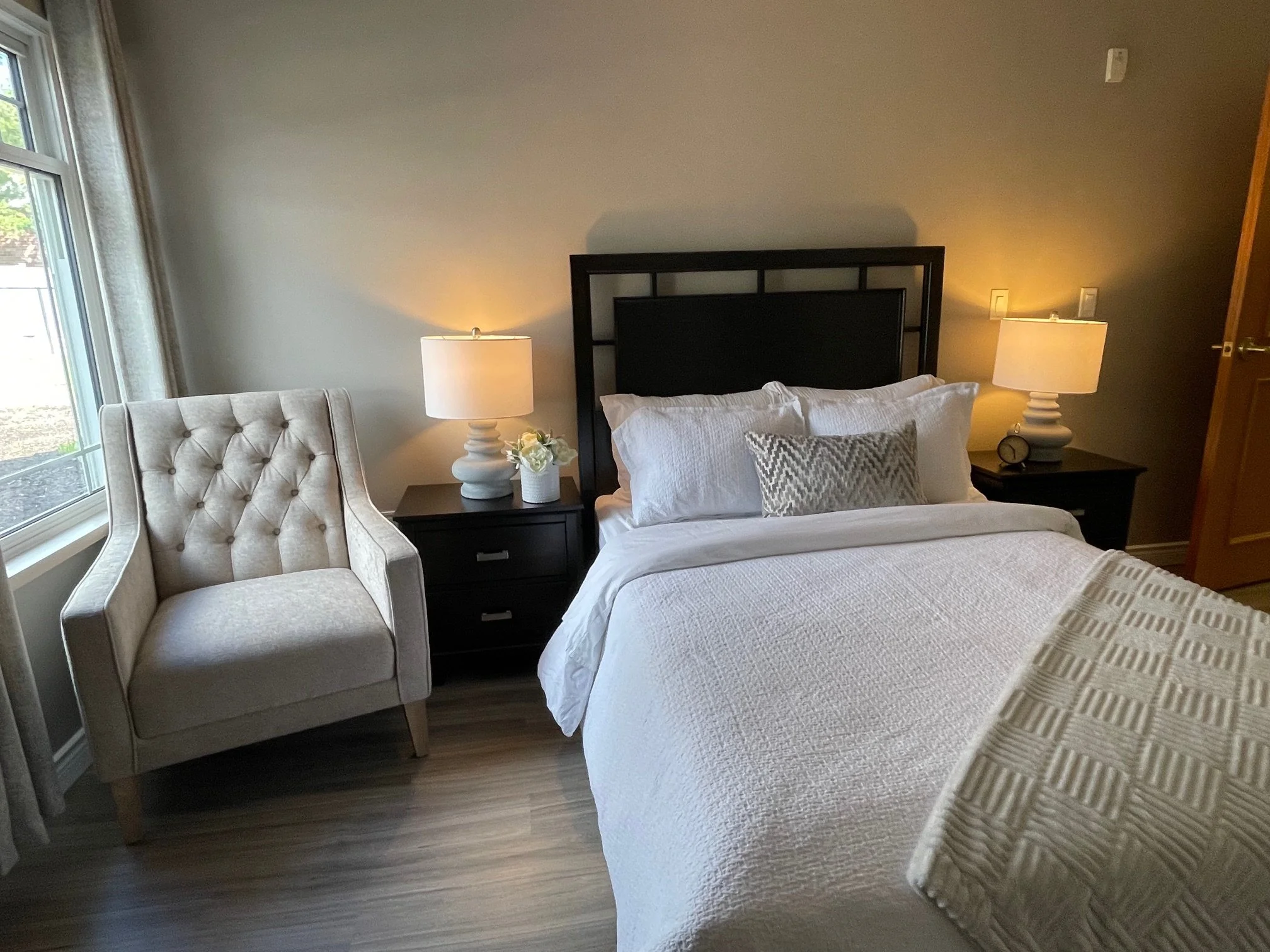 A cozy bedroom with a bed, white bedding, and a decorative pillow, flanked by two nightstands with matching lamps, a cream armchair near a window, and wood flooring.