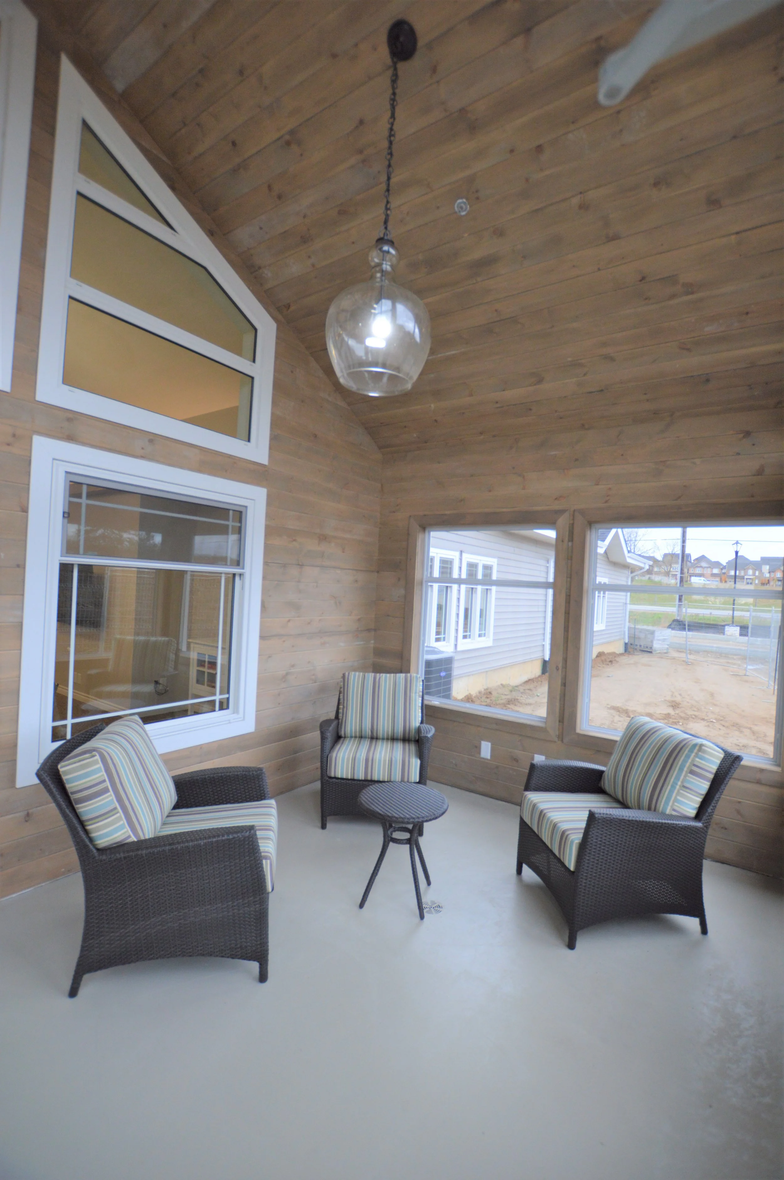 Indoor porch with three wicker chairs with striped cushions and a small round table, wooden walls and ceiling, two large windows, and a glass pendant light fixture hanging from the ceiling.