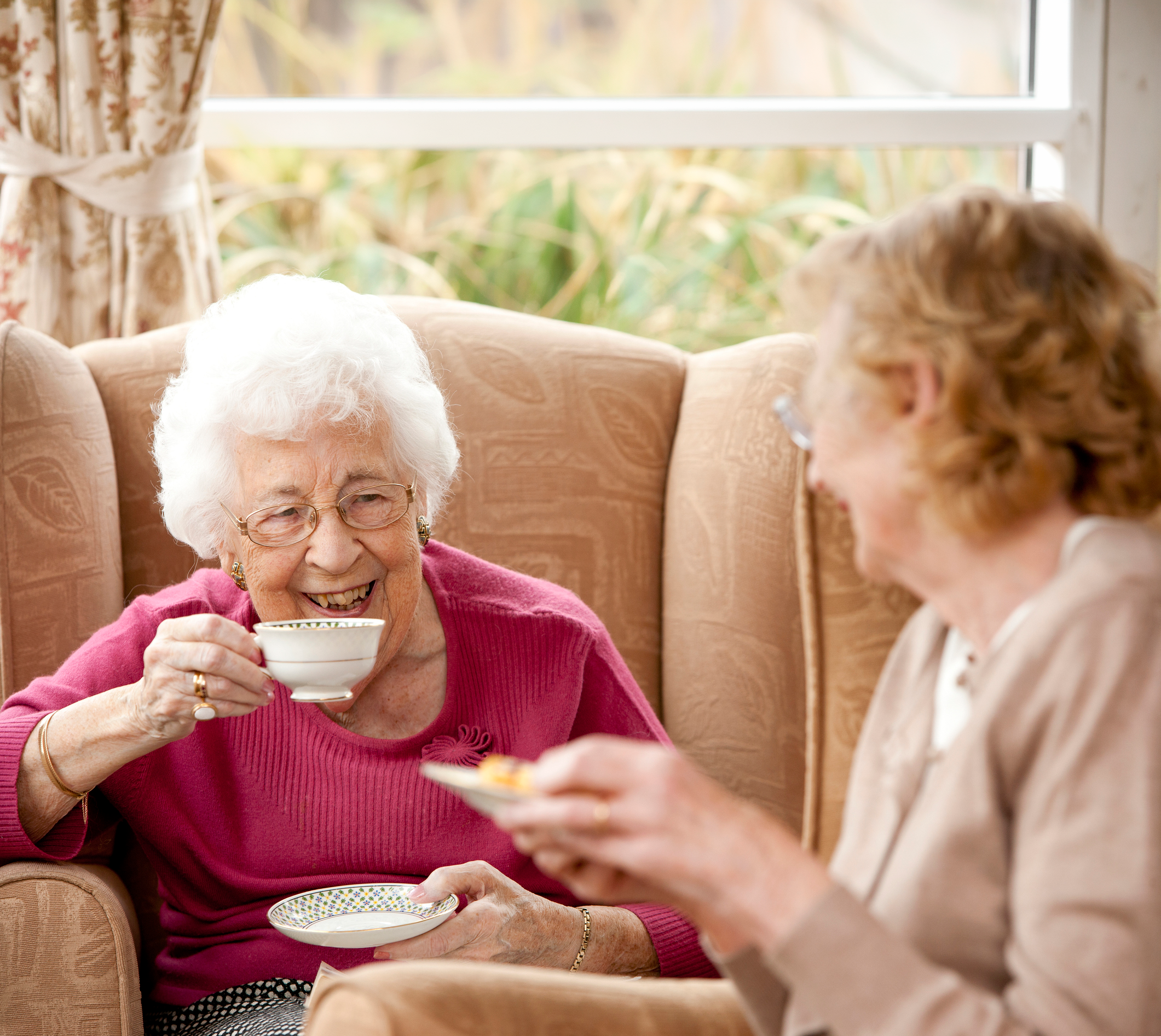 Two elderly women sitting on a sofa having tea and chatting, with a window and curtains in the background.
