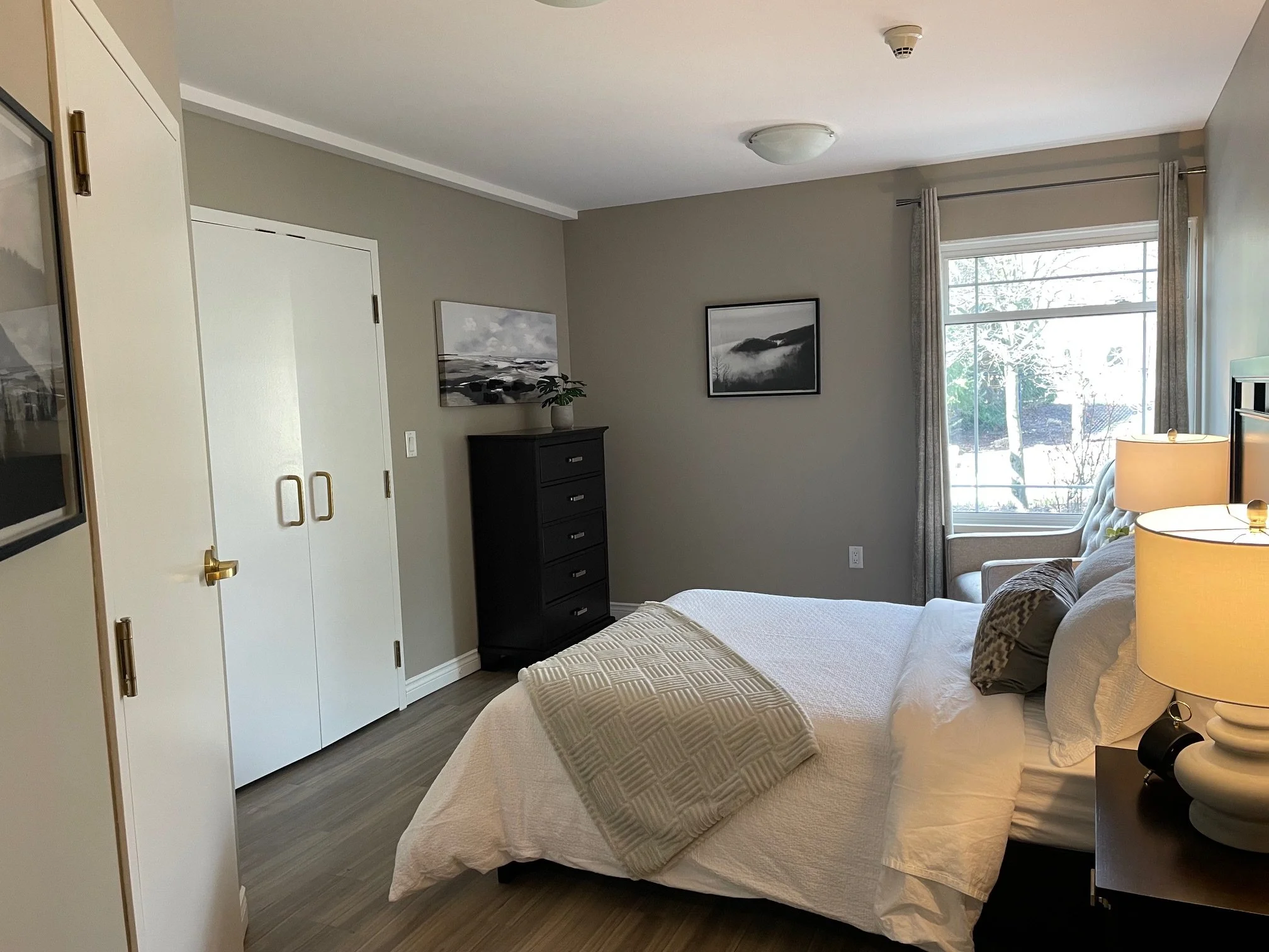A bedroom with beige walls, a white bed with pillows and beige blanket, a window with curtains, a black dresser, a reading chair by the window, picture frames on the wall, and a nightstand with a lamp.
