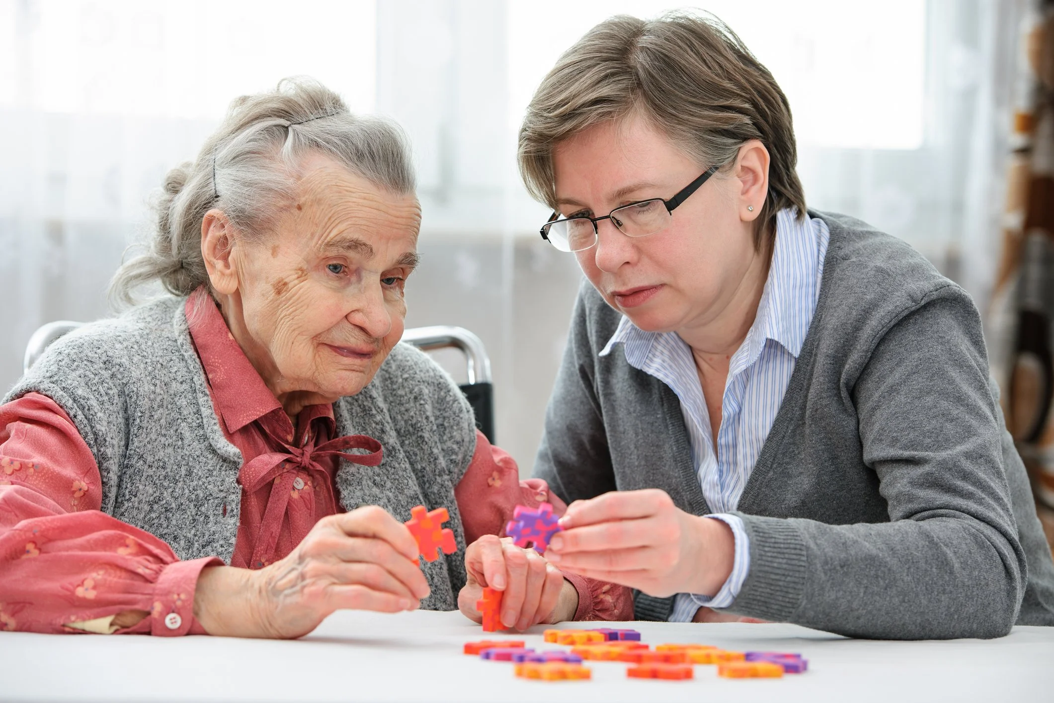 An elderly woman and a middle-aged woman sit at a table playing with colorful puzzle pieces. The elderly woman has gray hair and is wearing a pink blouse with a gray vest. The middle-aged woman has short hair, wears glasses, and a gray sweater over a blue collared shirt.