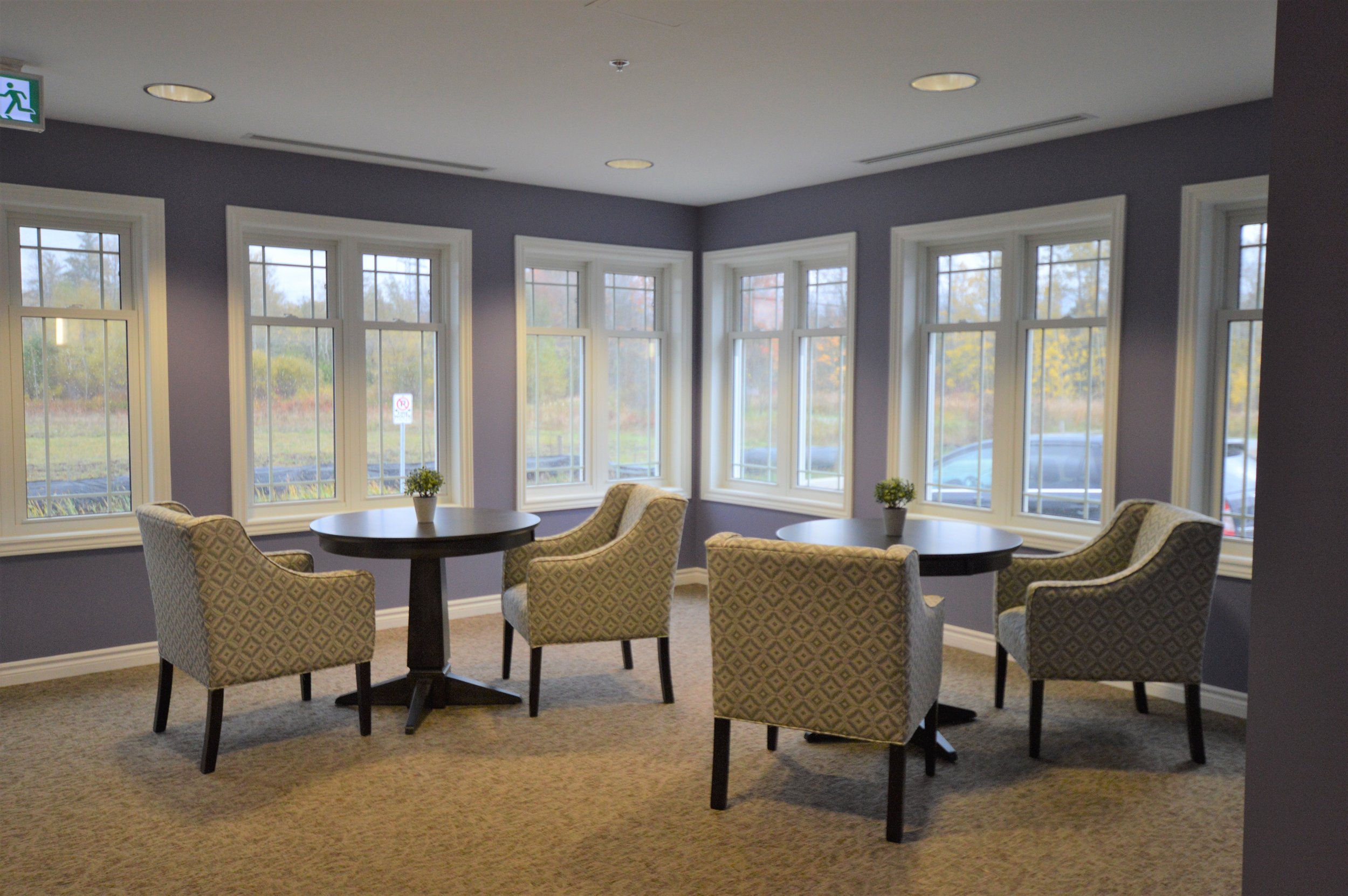 Sunlit corner of a room with seven large windows, two round tables, and four patterned armchairs with small potted plants on the tables.
