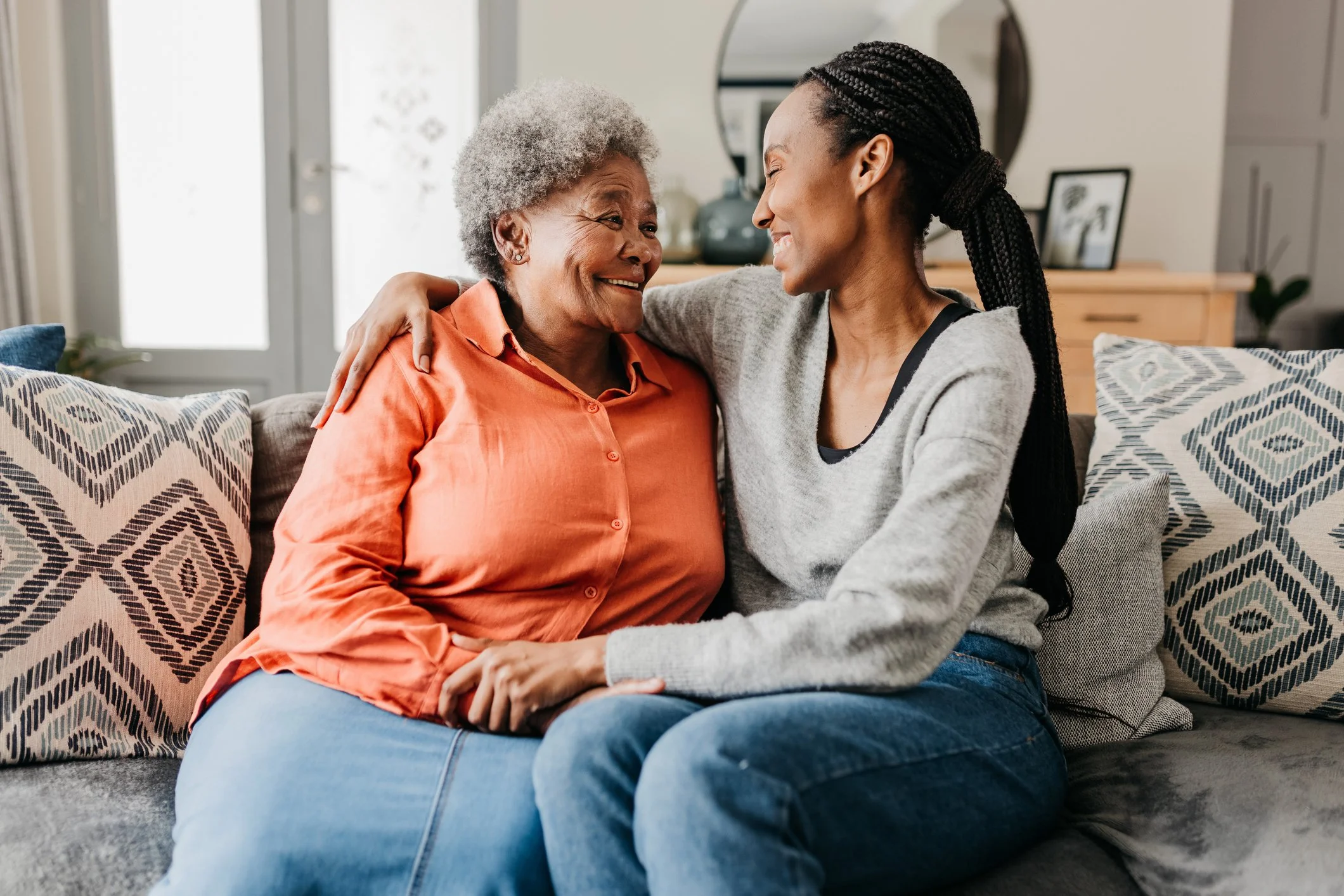 An elderly woman and a younger woman are sitting together on a sofa, smiling and looking at each other affectionately, with the younger woman embracing the older woman.