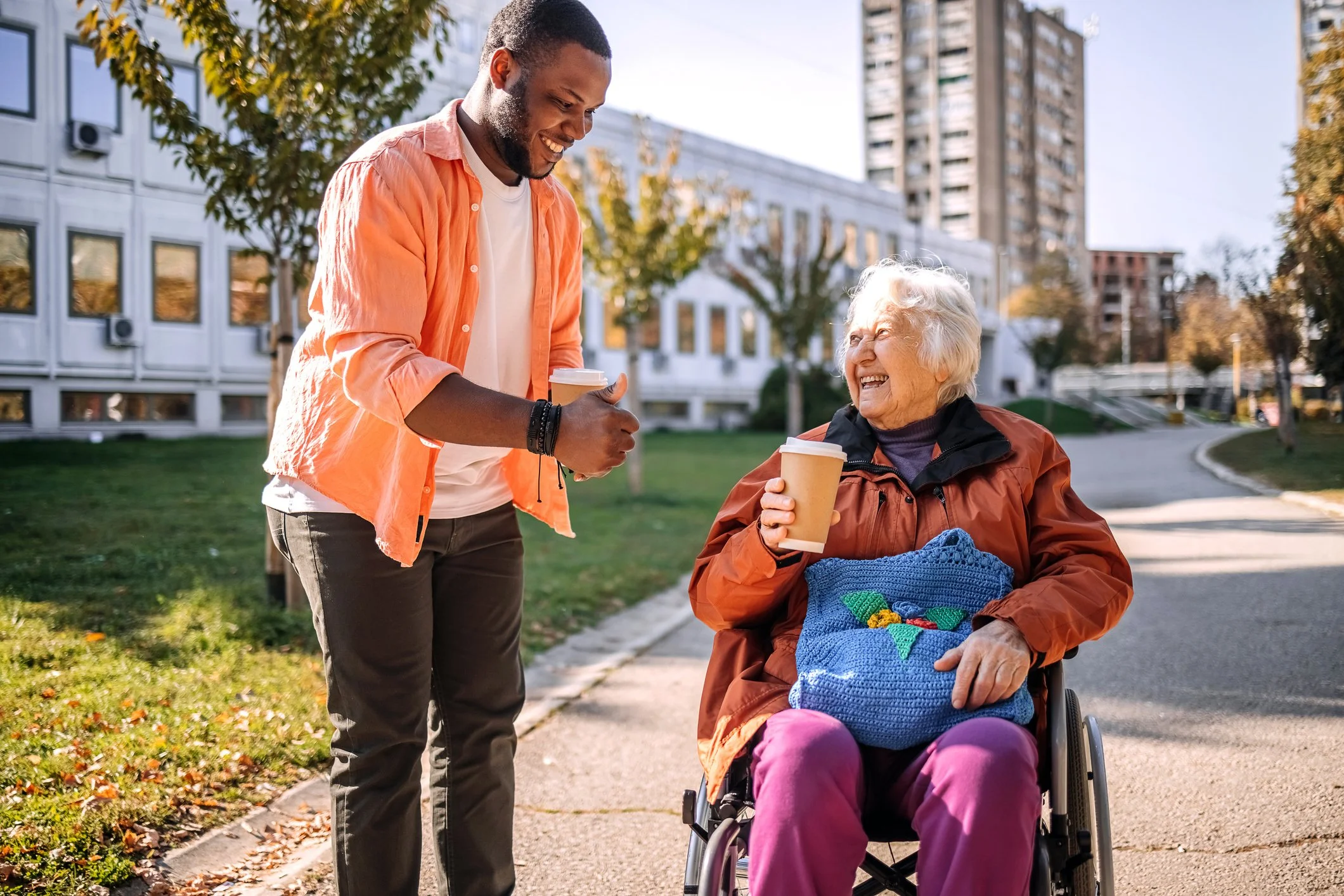 A young man in an orange shirt and gray pants handing a coffee cup to an elderly woman in a wheelchair, who is wearing a brown jacket and pink pants, smiling at each other outdoors on a sunny day.