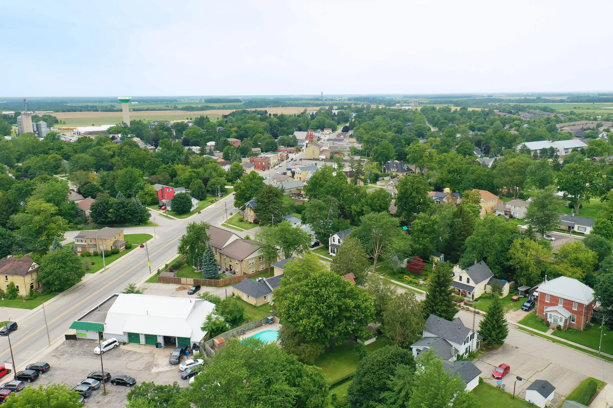 Aerial view of a small town with houses, trees, roads, and a water tower in the background under a light sky.