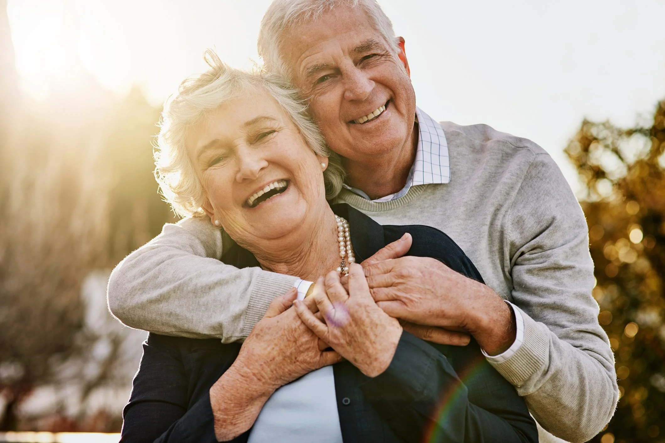 An elderly couple smiling and embracing outdoors during sunset, with the man hugging the woman from behind, both appearing happy.