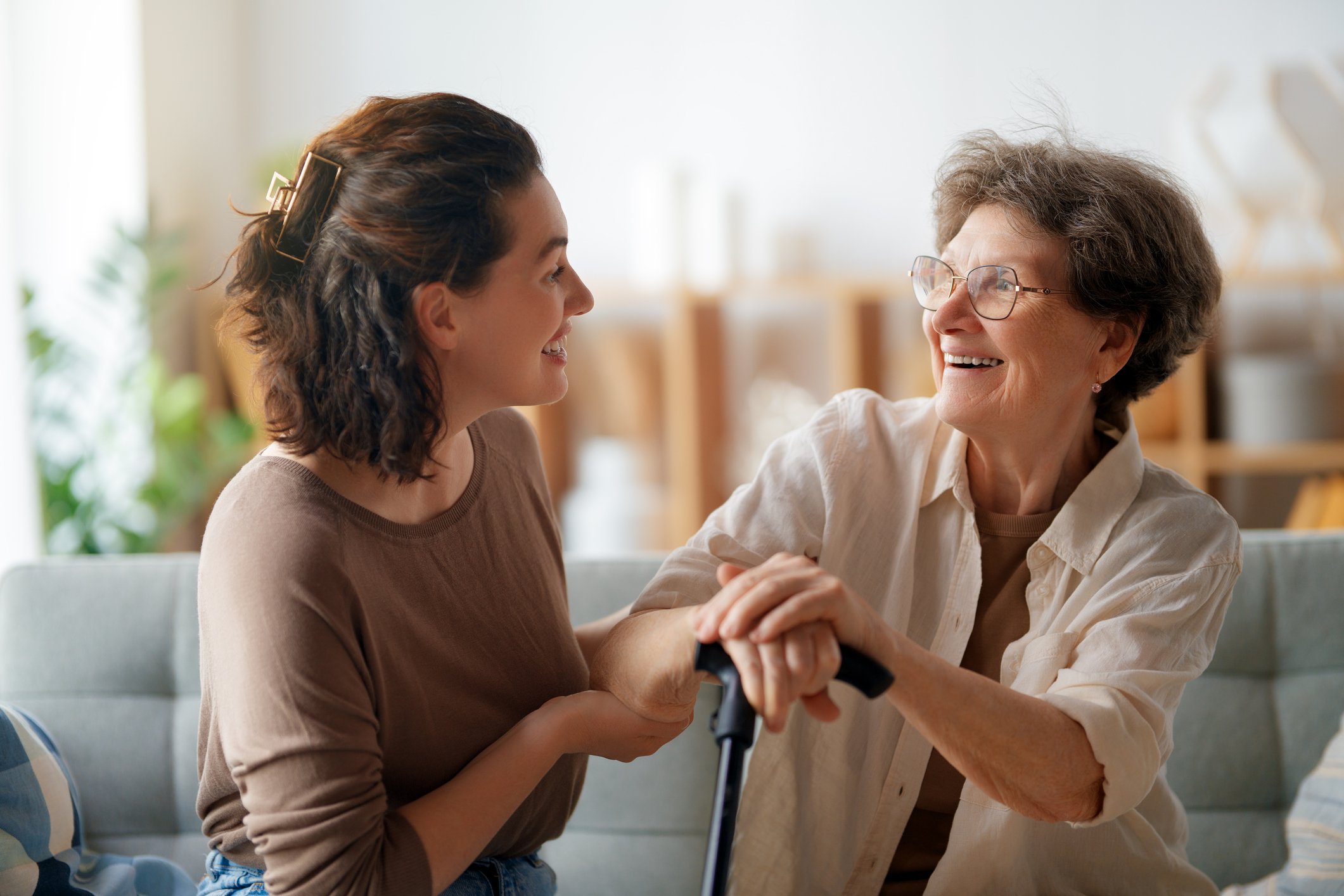 A young woman and an older woman are sitting on a couch, smiling and holding hands. The older woman has short gray hair, glasses, and is holding a walking cane. They are in a bright, cozy living room.