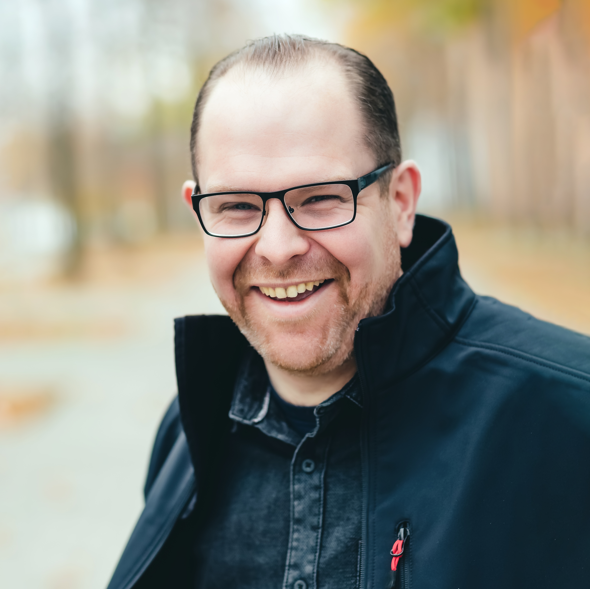 A smiling man wearing glasses, a black jacket, and a dark shirt, outdoors with blurred trees in the background.