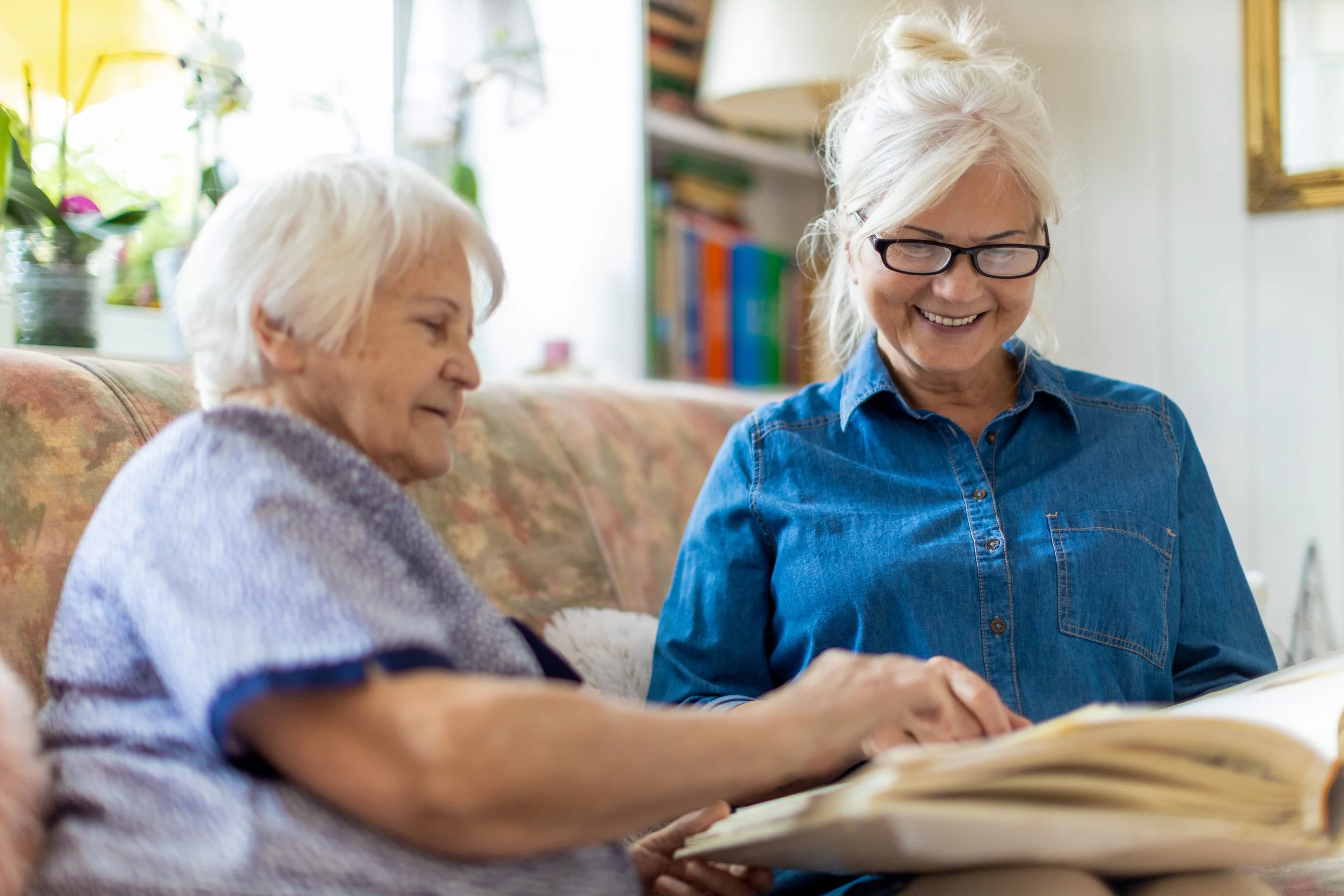 Two women, one elderly with white hair wrapped in a blanket, and a middle-aged woman with glasses and blonde hair in a bun, sitting on a patterned couch, looking at a large photo album together in a cozy, well-lit living room.