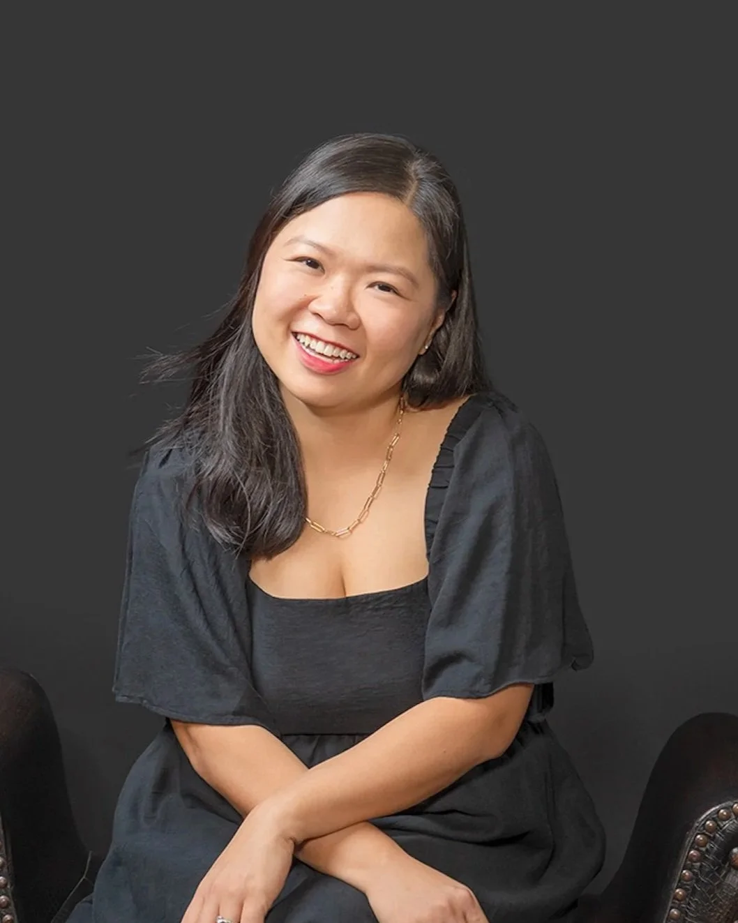 A woman with long dark hair smiling, wearing a black dress and gold jewelry, sitting against a dark background.