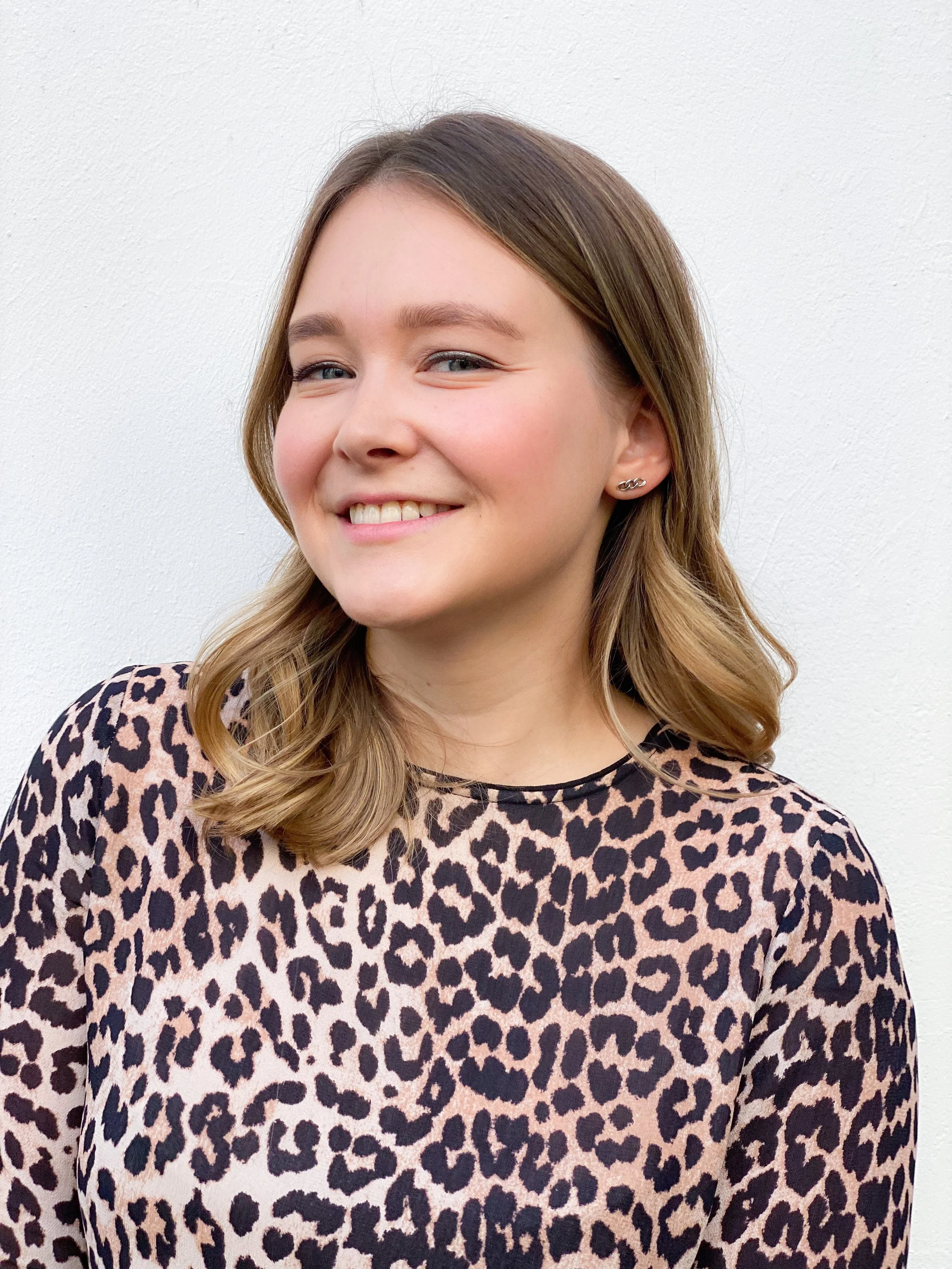 Junge Frau mit braunen, gewellten Haaren trägt ein leopardmusterndes Oberteil und lächelt vor weißer Wand.