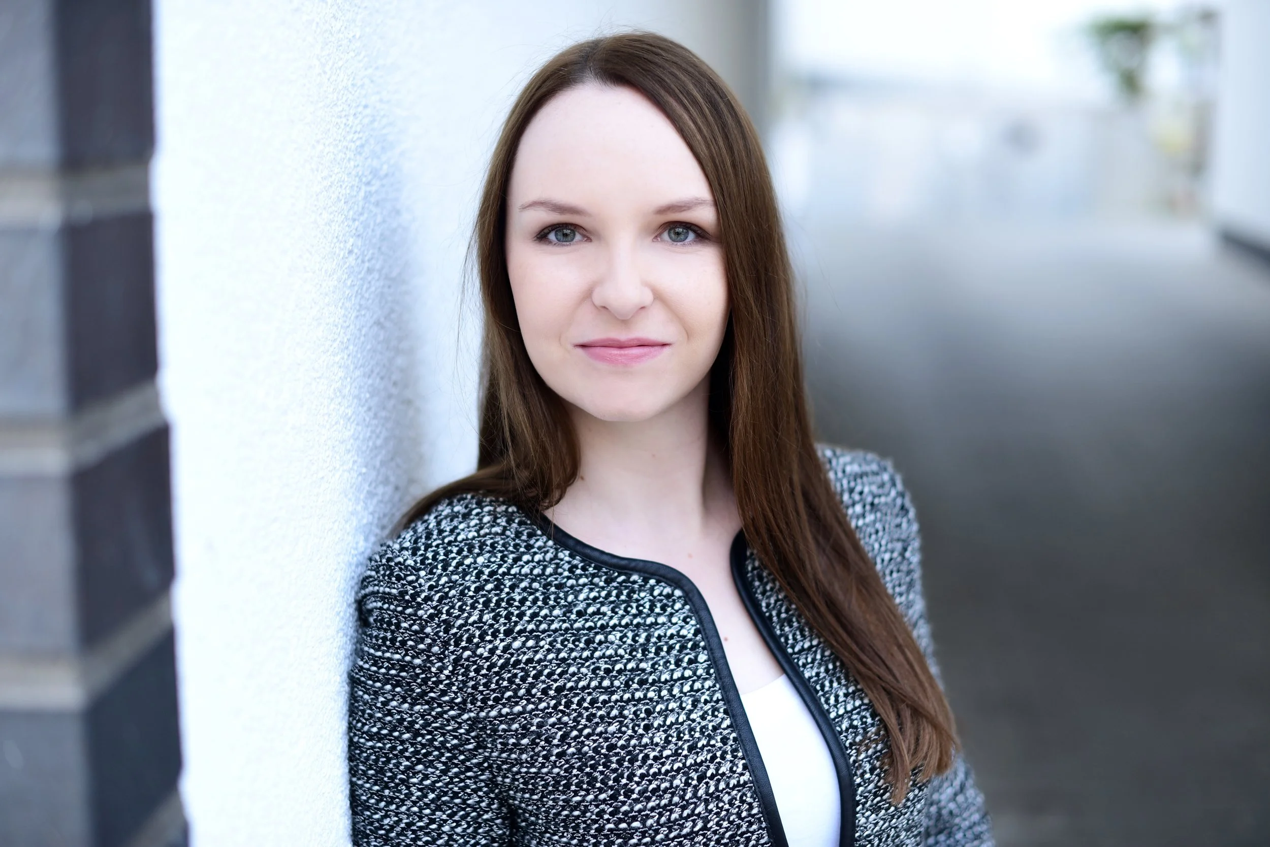 Junge Frau mit langen braunen Haaren und blauen Augen, trägt eine schwarze und weiße Jacke, steht an einer Außenwand, blickt freundlich in die Kamera.