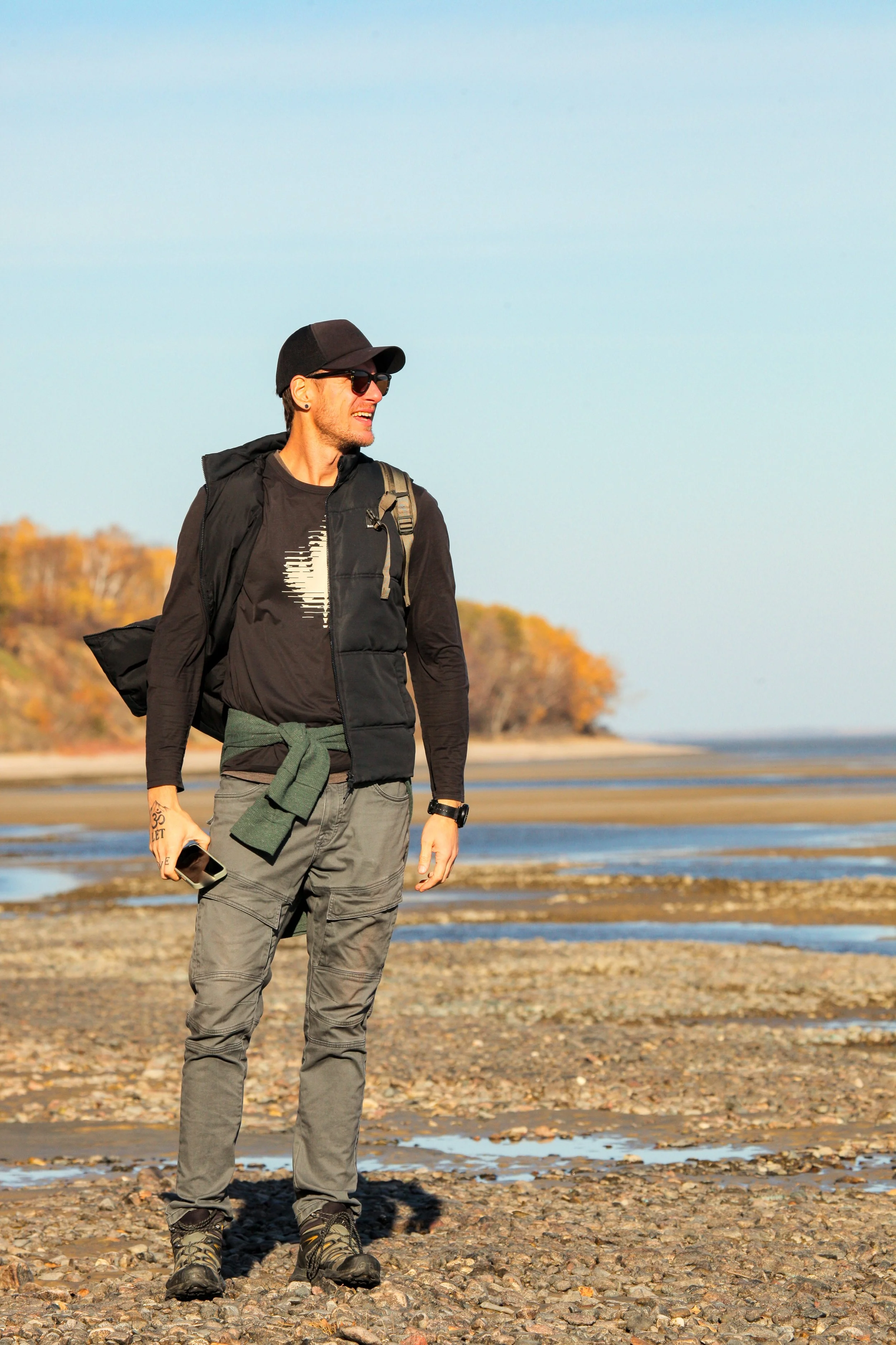 A man with dark hair, sunglasses, and tattoos wearing a black jacket over a t-shirt and gray cargo pants, walking on a pebble beach with trees and water in the background under a clear sky.
