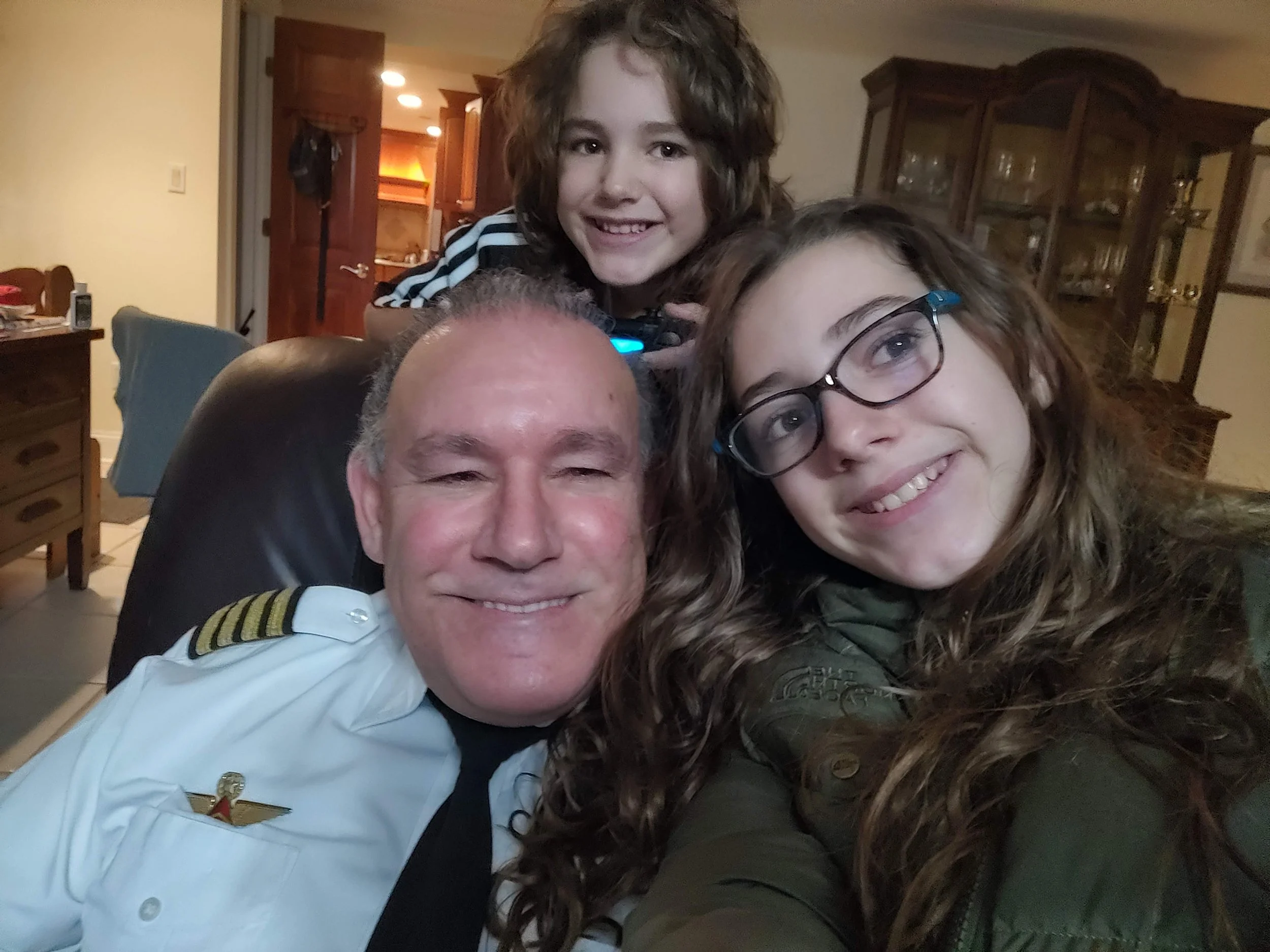 Family of four smiling and taking a selfie in a cozy living room with wooden furniture and warm lighting.