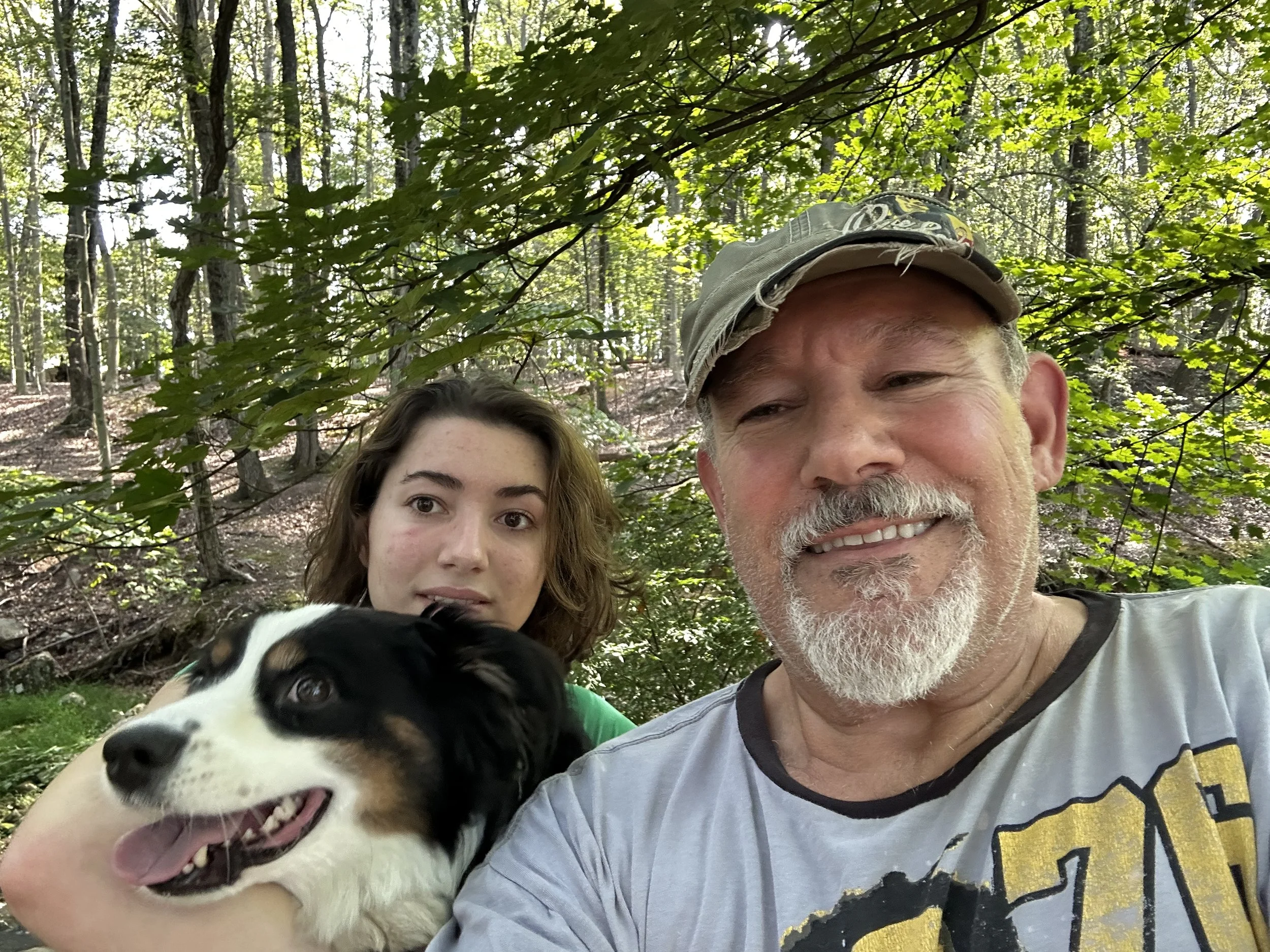 Two people, an older man wearing a cap and a young woman, are posing for a selfie in a lush green forest. A black and white puppy is in the foreground.