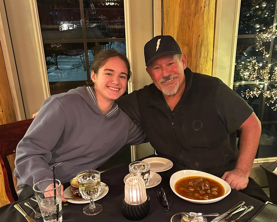 A young woman and an older man sitting at a dining table, smiling and posing for the photo. The table has glasses of water, a bowl of soup, and a candle. The background shows a window with a winter scene outside.