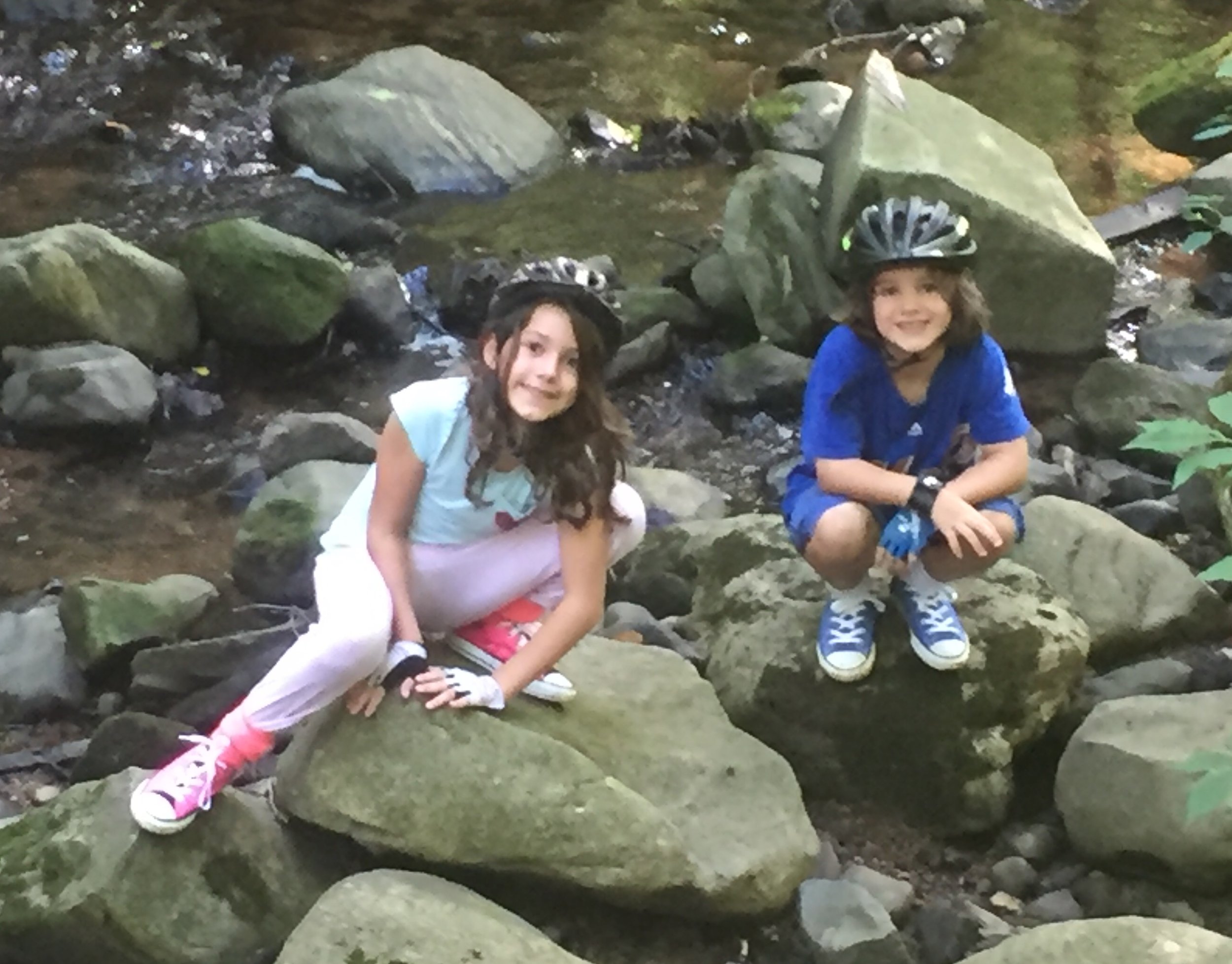 Two children wearing bicycle helmets and gloves are sitting on rocks by a creek, smiling at the camera.
