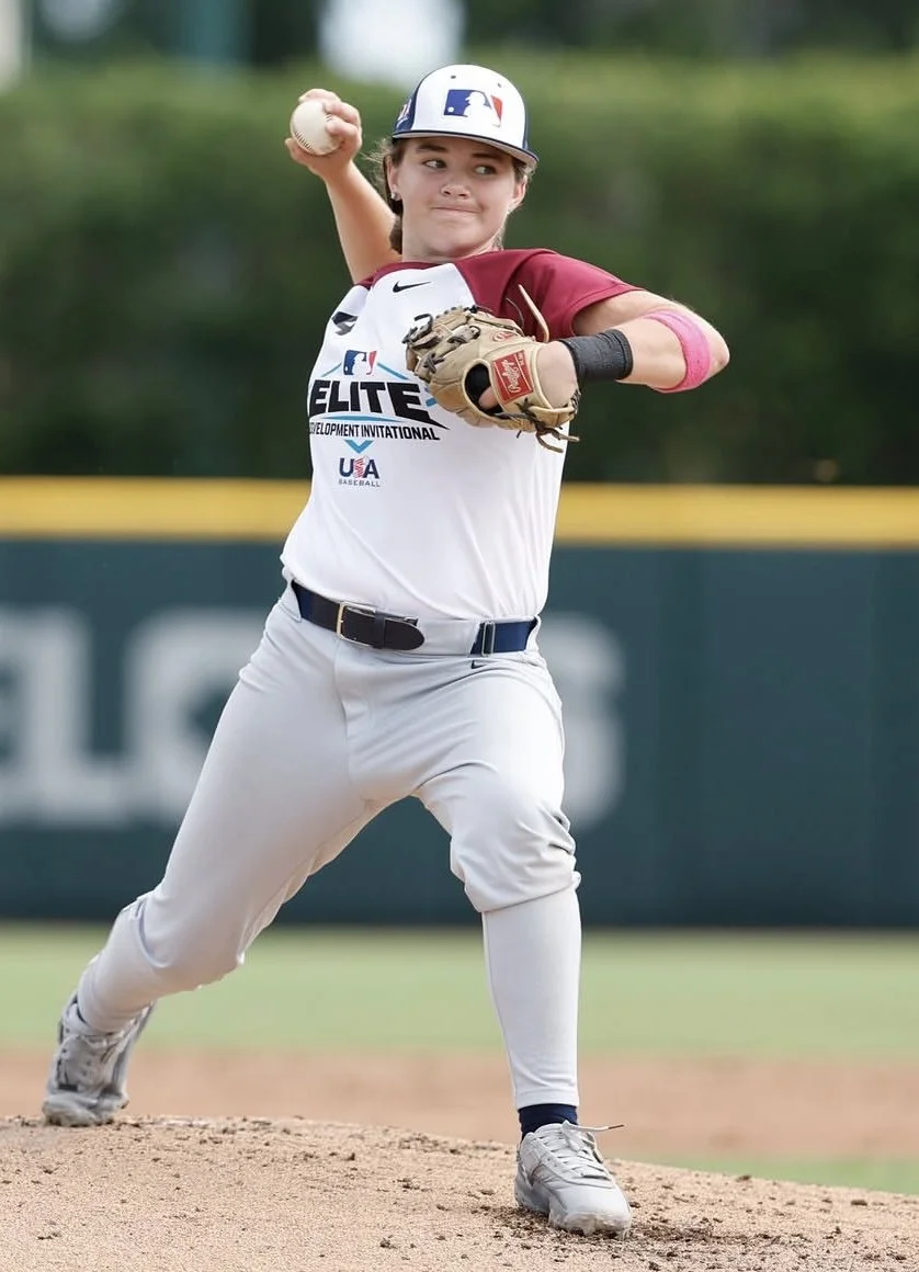 A young girl in baseball uniform throwing a baseball on a baseball field.