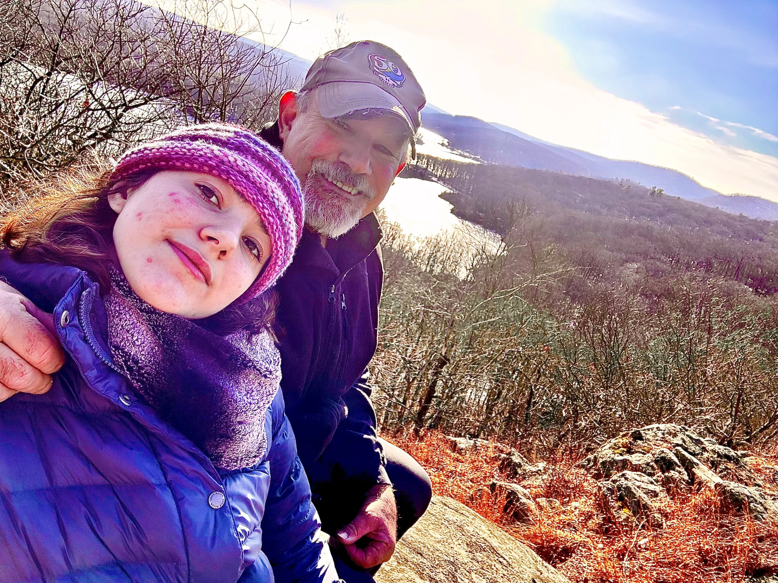 A young girl and an older man taking a selfie outdoors with a mountain and a river in the background.