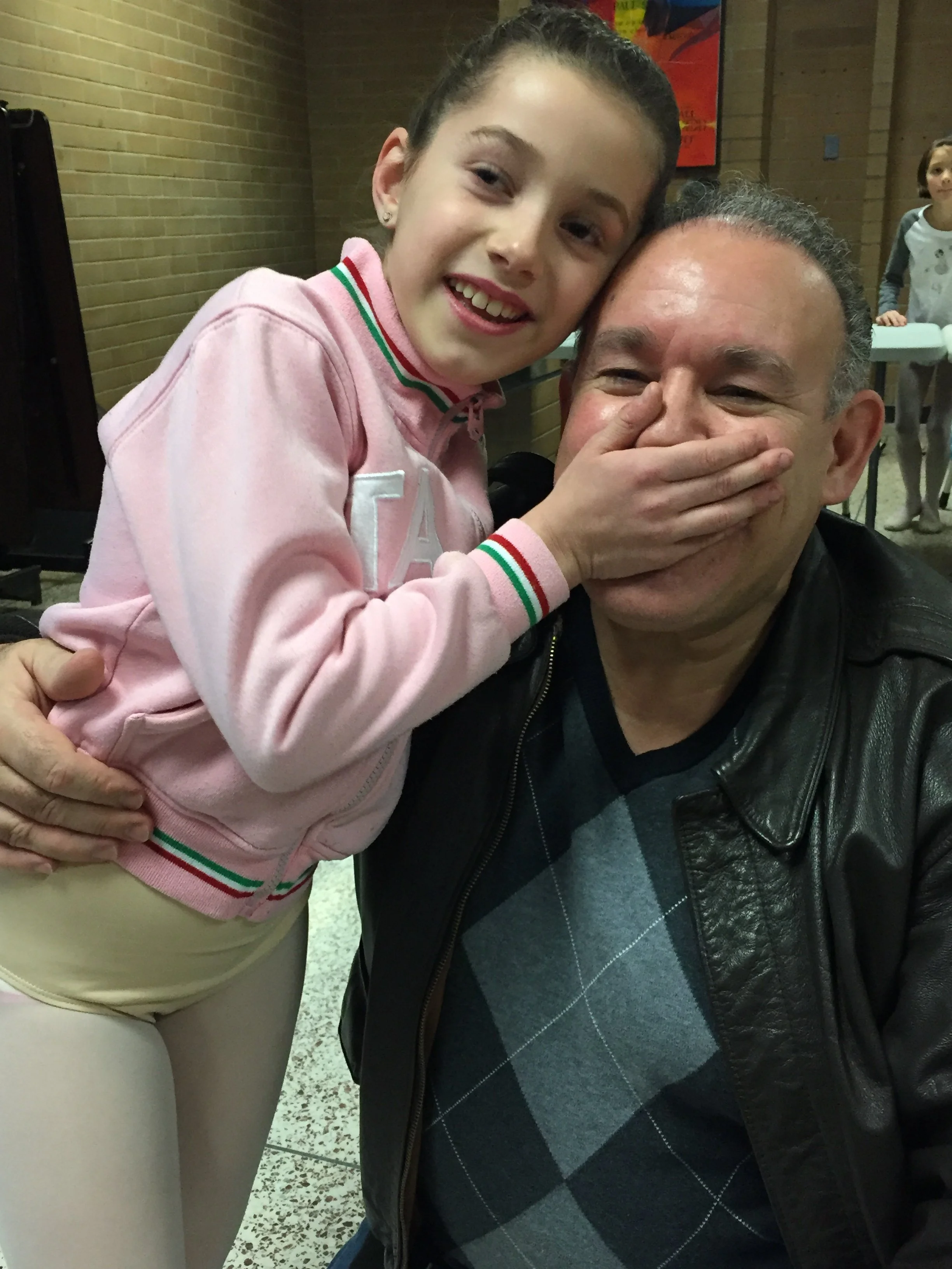 A young girl covering her mouth with her hand, smiling, standing close to a man in a black leather jacket, both smiling. The girl is wearing a pink jacket with a tricolor trim and beige pants. The background shows a brick wall and another woman in the distance.