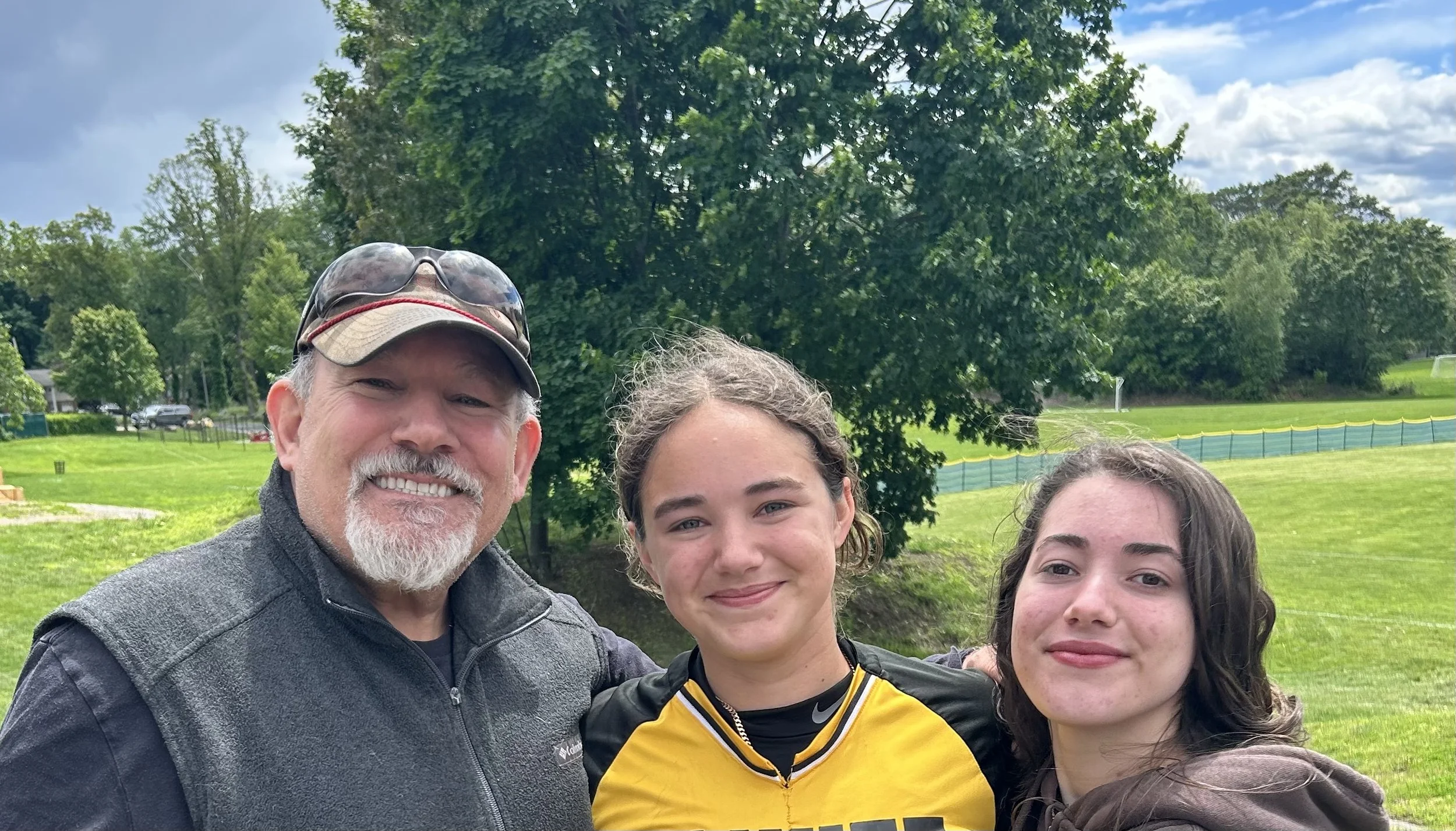 A group of three people, including an older man with glasses and a beard, and two young women, smiling outdoors with trees and a grassy field in the background.