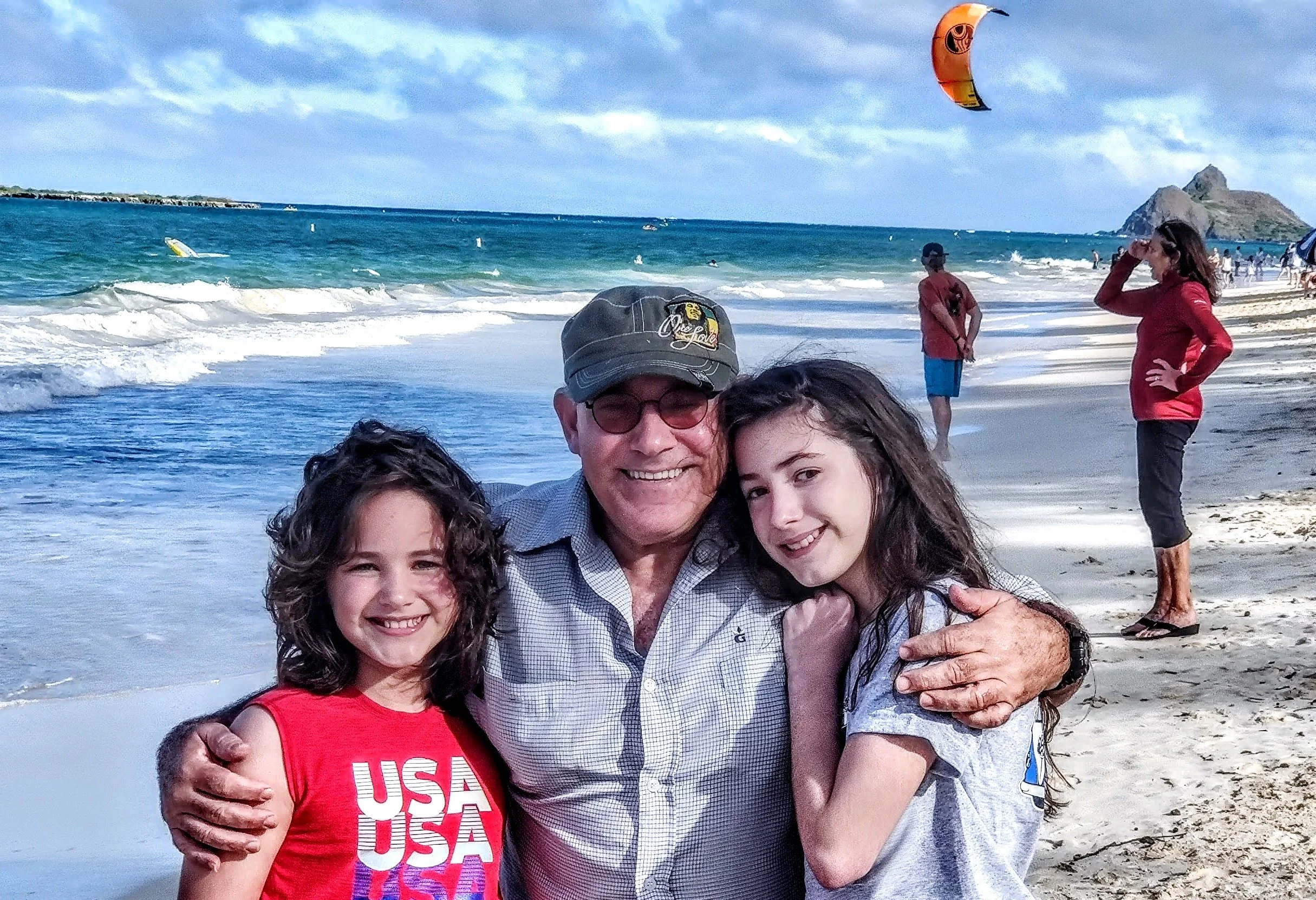 A family of five enjoying a day at the beach, smiling and hugging each other, with ocean waves, a partly cloudy sky, and some people flying kites in the background.