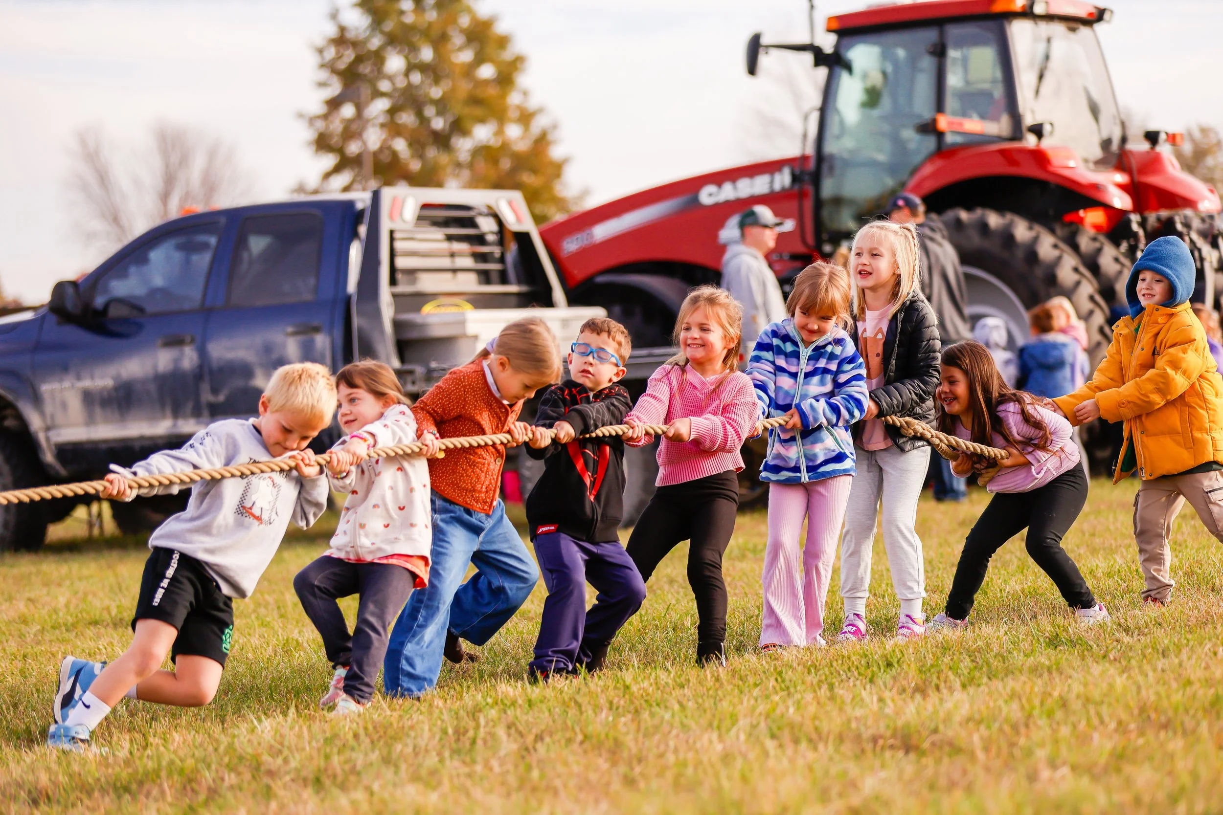 Children playing tug-of-war outdoors on grassy field with farm equipment and a red tractor in the background.