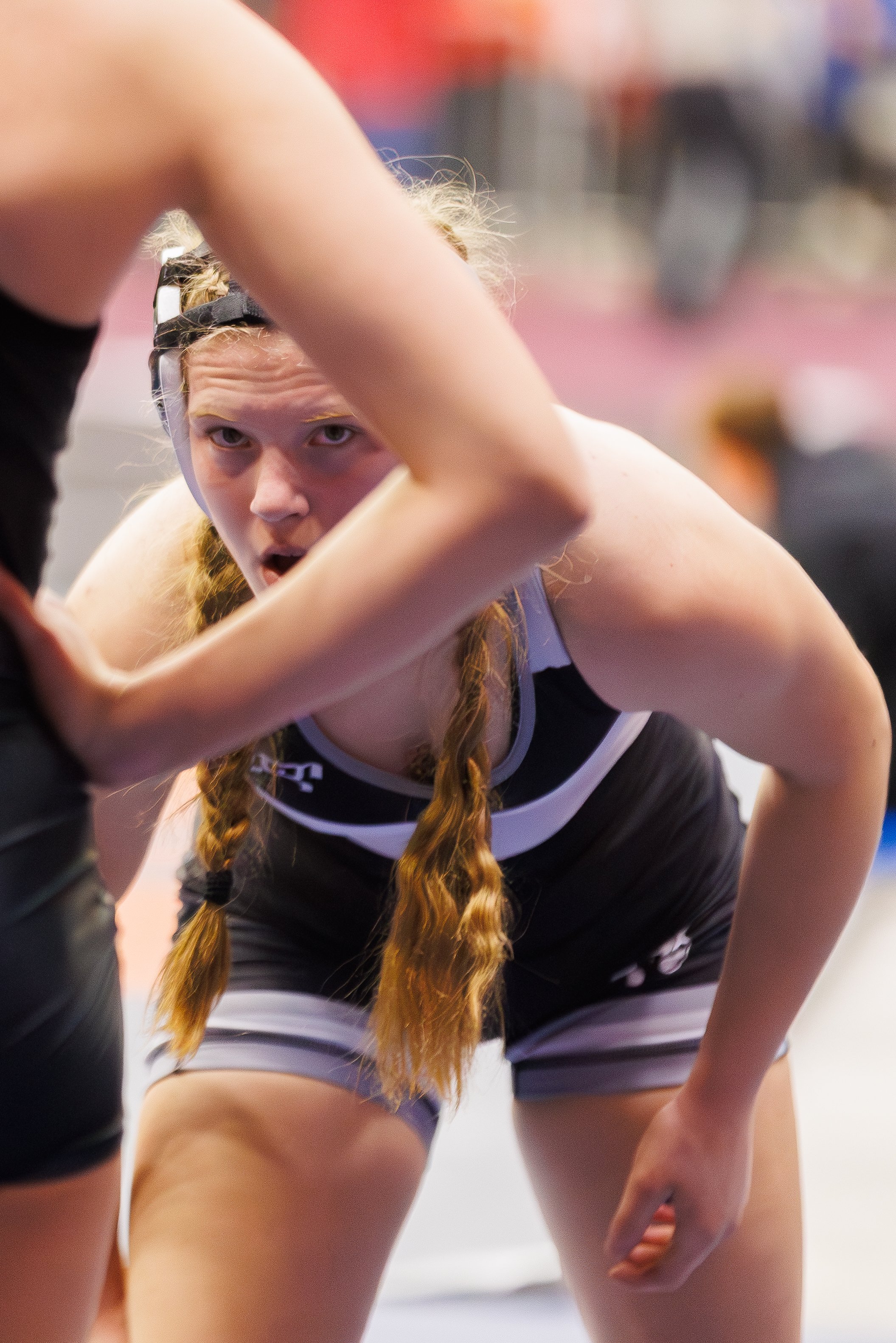 A female wrestler with long red hair in braids, wearing a black and white singlet, has an intense expression while engaged in a wrestling match, crouched in a defensive stance.