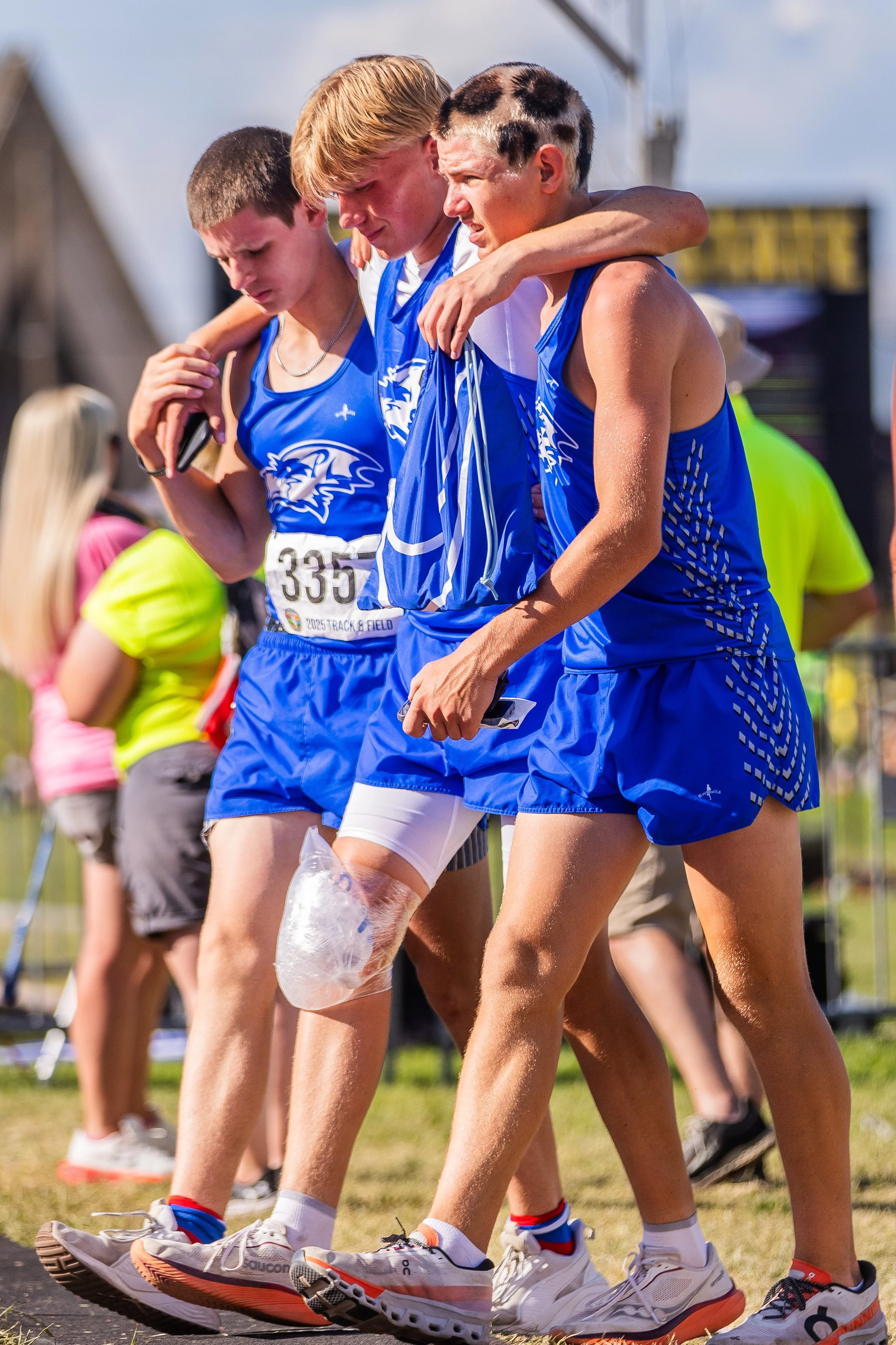 Three young male athletes in blue track uniforms with white and blue design, standing together, their arms around each other's shoulders, after a race, with a bandage on one runner's knee, and other runners and spectators in the background.