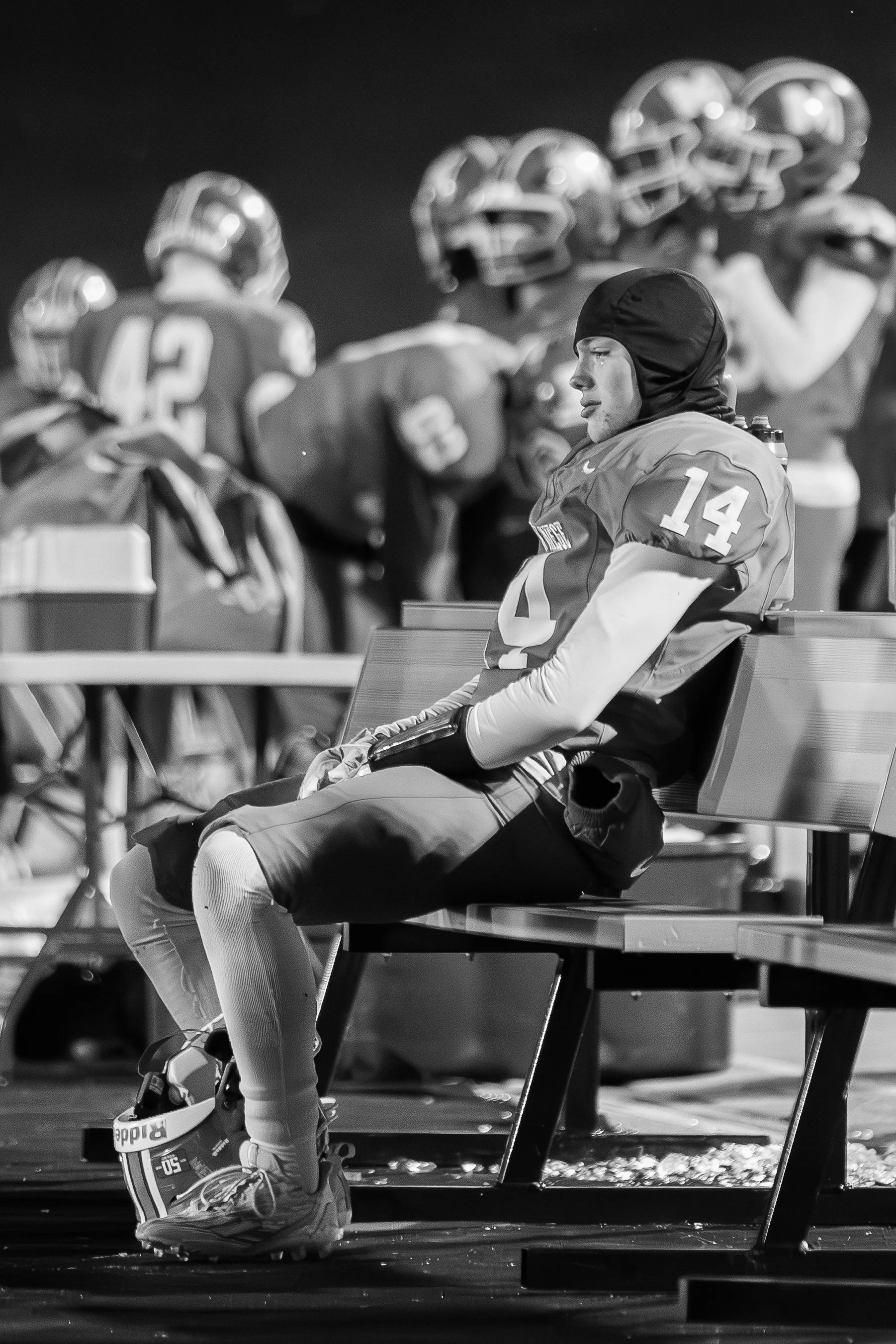 Black and white photo of a football player sitting on a bench with a hoodie on, wearing a jersey with the number 14. In the background, other players are standing and preparing on the sideline.