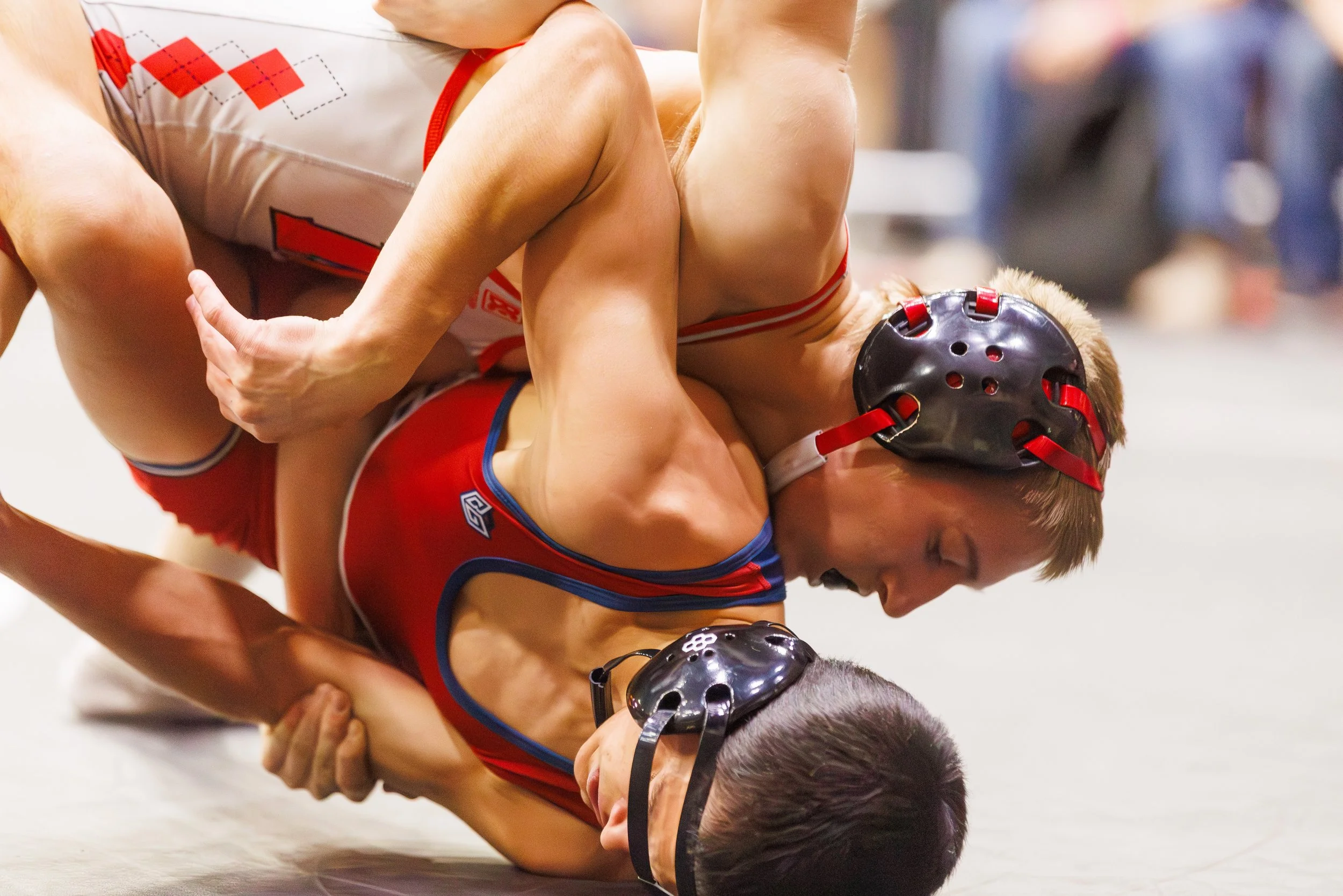 Two female wrestlers competing on a mat, wearing protective headgear, one in a red singlet and the other in a white singlet with red accents, engaged in a grappling hold.