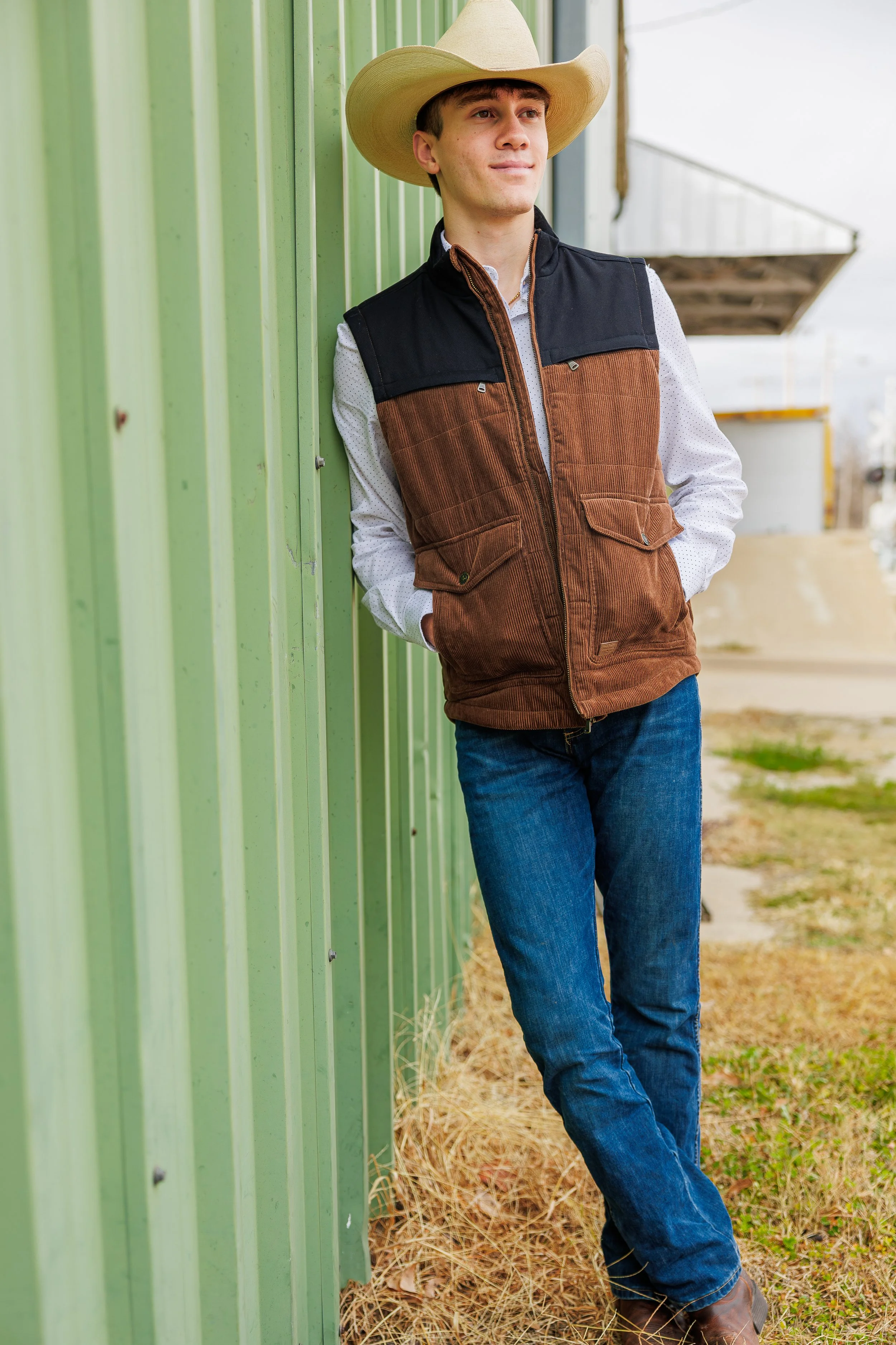 Young man in cowboy hat leaning against green metal building, wearing vest, white shirt, jeans, and boots.