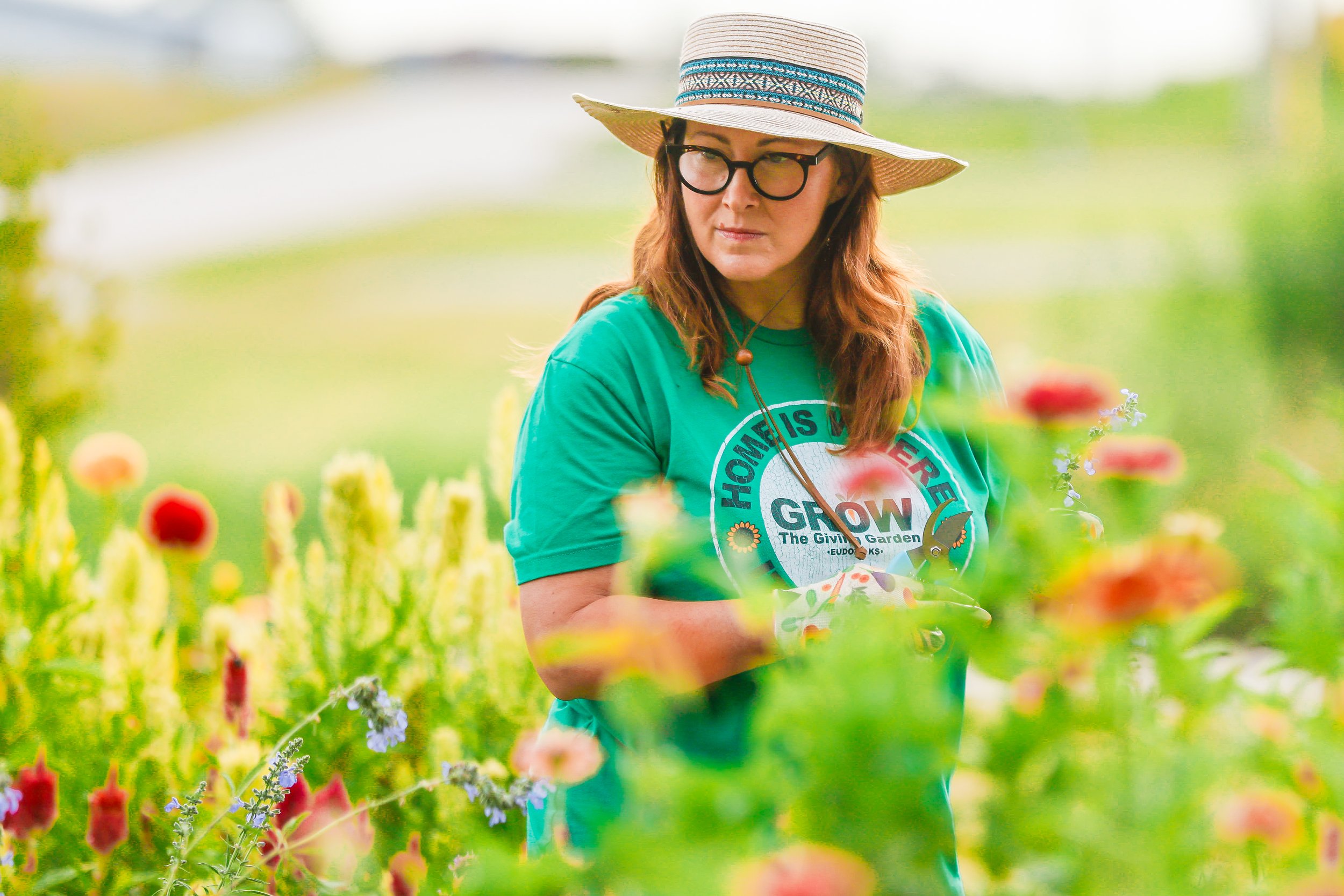 A woman wearing a wide-brimmed hat, glasses, and a teal T-shirt with a gardening logo, standing in a lush garden with blooming flowers.